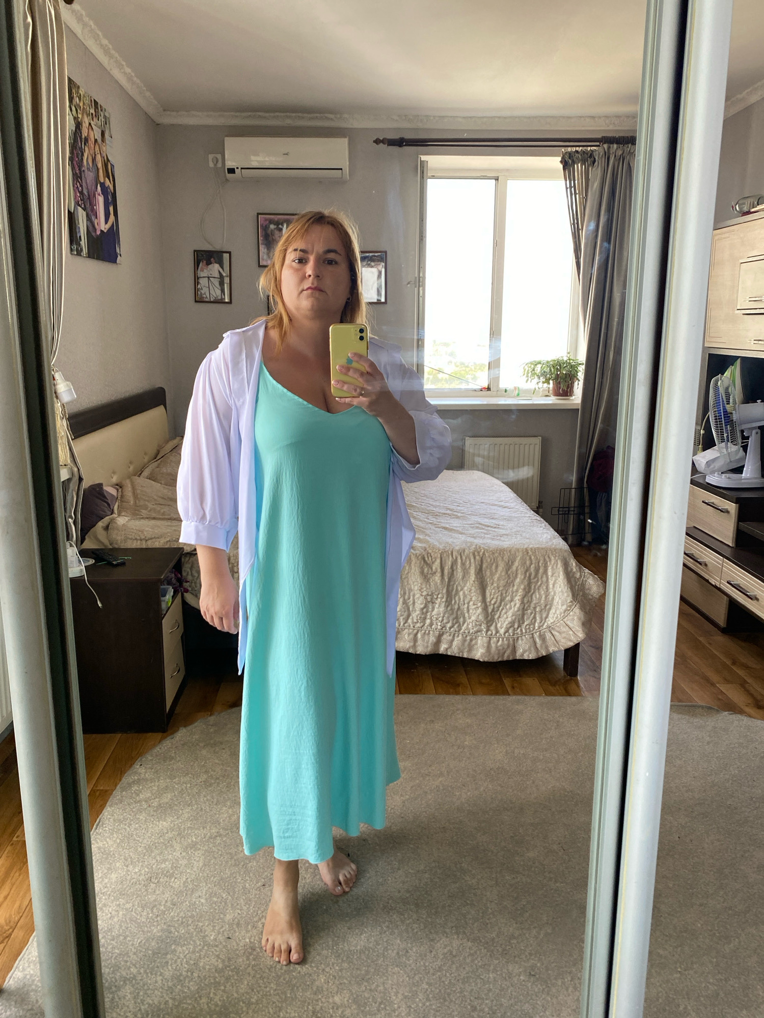 A full-body mirror selfie shows a woman with blonde hair, wearing a light blue spaghetti strap dress and a white sheer kimono. She is holding a yellow smartphone in her right hand, taking the photo. The background is a bedroom with a bed, nightstand, dresser with a fan, and a window. The room appears to be in an apartment in Chornomorsk, Ukraine, as indicated by the overall decor and style. The woman is standing on a rug, barefoot.
