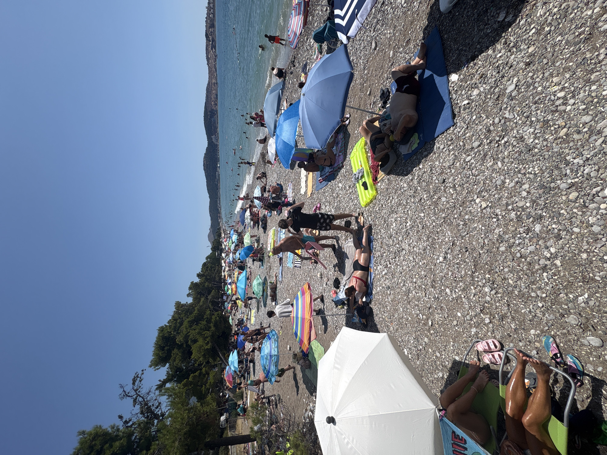 This is a wide-angle, eye-level shot of a crowded pebble beach in Bar, Montenegro on a sunny day. The sea is to the right, with several people swimming and wading in the blue water. The beach is filled with sunbathers, many under colorful umbrellas, lounging on towels and mats. Some individuals are standing, while others are lying down. A prominent white umbrella is in the foreground on the left, and a few people are relaxing underneath it. The background shows a tree-lined coast with hills and buildings in the distance.