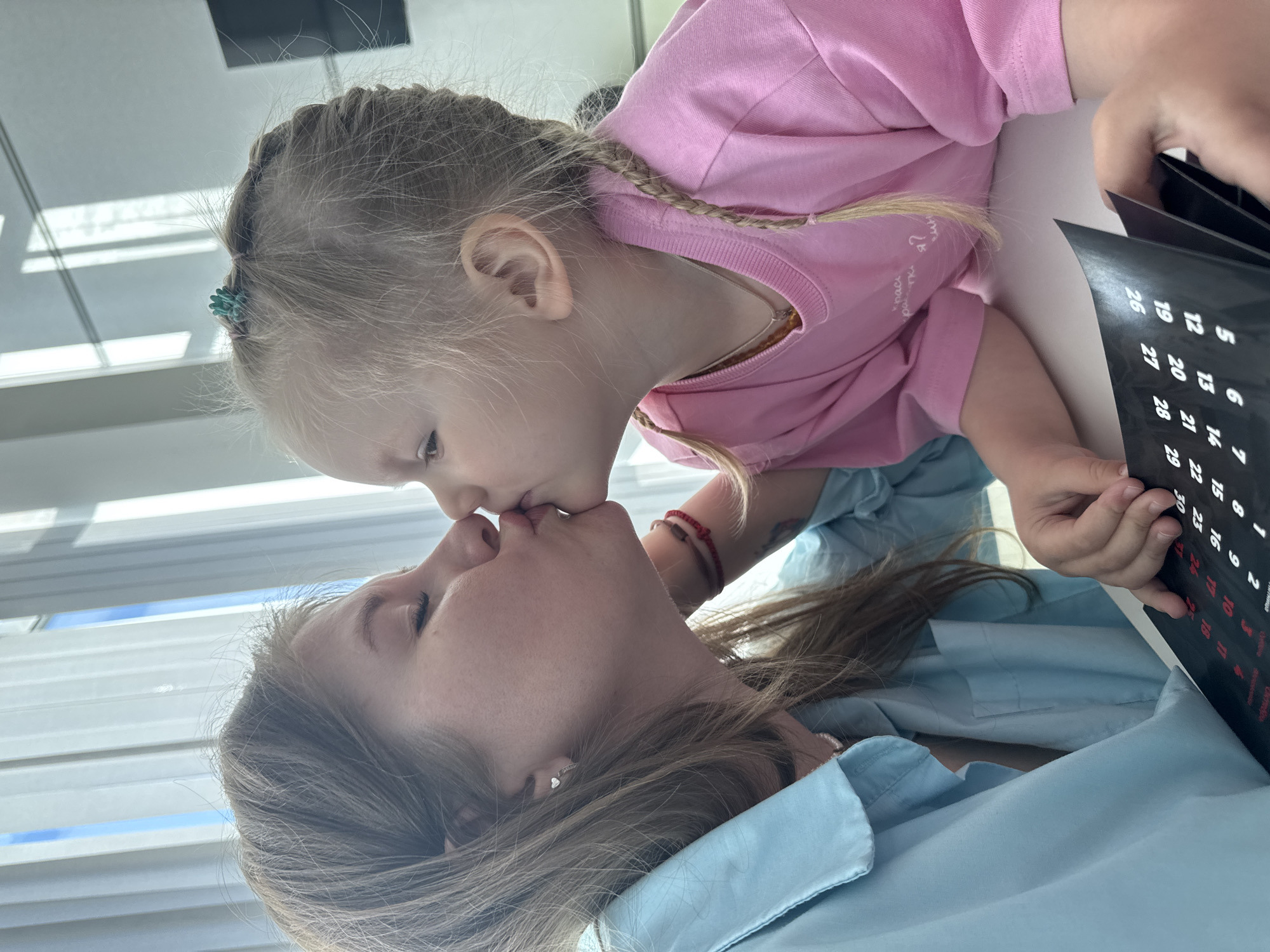 This rotated indoor shot, set in Perm, Russia, captures an affectionate moment between a young girl and an adult woman during daylight. Both subjects are leaning over a white surface, possibly a table, with natural light filtering through window blinds in the background.
The young girl, with blonde hair styled in multiple braids, one of which is adorned with a small turquoise hair clip, wears a pink long-sleeved shirt. Partially visible white text on her shirt reads "рассказать я" (to tell I/me). She has a thin necklace and a red string bracelet on her left wrist. She is leaning in to gently kiss the adult woman on the lips, conveying tenderness and connection.
The adult woman, with brown hair, wears a light blue collared shirt and a small heart-shaped earring in her right ear. Her eyes are closed, indicating she is receiving the kiss.
In the girl's hands, she holds a black object, likely a calendar or planner, which displays numerous white and red numbers. Visible numbers include "26, 19, 12, 5, 27, 20, 13, 6, 1, 28, 21, 14, 7, 2, 29, 22, 15, 8, 3, 30, 23, 16, 9, 4, 24, 17, 10". The overall mood of the scene is intimate and loving.