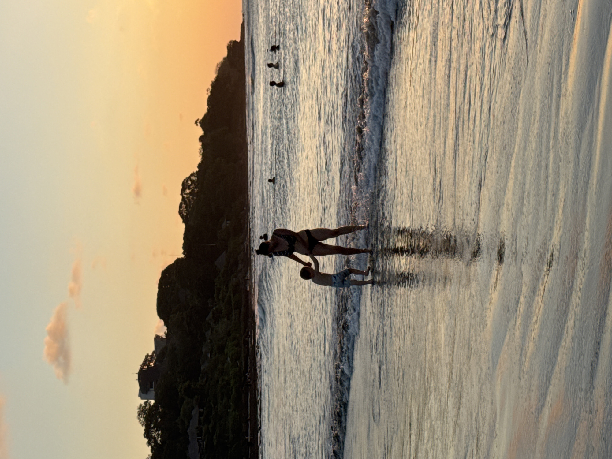 This media file depicts a serene beach scene at sunset in Kuta Selatan, Indonesia. The subjects are a woman and a child, silhouetted against the glowing sky and the reflective water. They appear to be engaged in a gentle interaction on the wet sand at the water's edge. The woman, wearing a bikini, is holding the child's hand. The child, in swim trunks, stands facing away from the viewer. Several other silhouetted figures are visible further out in the water, suggesting others are also enjoying the beach.
The time of day is clearly sunset, indicated by the warm, gradient colors in the sky, transitioning from a soft orange and pink to a lighter yellow and pale blue. Wispy clouds add texture to the sky. The weather appears calm, with gentle waves lapping at the shore, creating rippling patterns on the sand. The light from the setting sun casts long shadows and highlights the textures of the wet sand and the water.
In the background, a lush, dark green hillside covered in trees slopes down towards the beach. Some structures are partially visible amongst the foliage, suggesting a developed area or resort along the coast. The overall mood is peaceful and tranquil, capturing a moment of connection and enjoyment between the woman and child, as well as the natural beauty of the tropical sunset. There is no visible text in the media file.