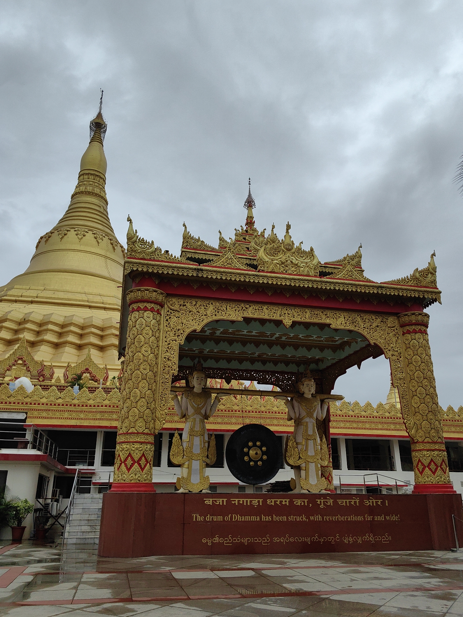 The image shows the Global Vipassana Pagoda in Gorai, Mumbai, India. The pagoda is a prominent golden structure with intricate detailing and a tall spire. In the foreground, there is an ornate archway supported by statues. A large gong is visible within the archway. Text is inscribed on a red base in front of the archway, with the English translation "The drum of Dhamma has been struck, with reverberations far and wide!". The ground in front of the structure is wet, indicating recent rain. The sky is overcast.