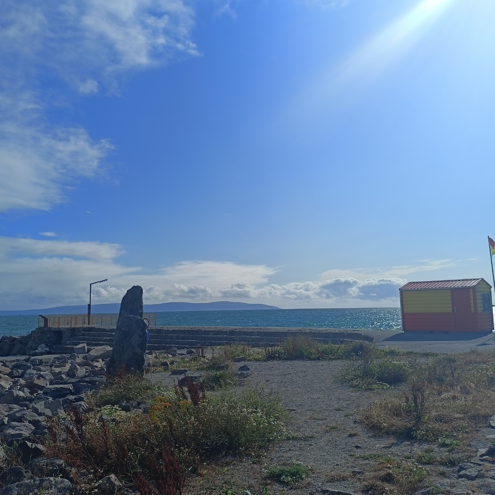 This is a photograph of a coastal scene in Galway, Ireland. The image features a bright blue sky with scattered white clouds, and the sun is shining brightly, creating a lens flare in the upper right quadrant. In the foreground, there is a path made of gravel and scattered rocks, with some dry vegetation and weeds growing.
To the left, a large, dark, pointed stone stands prominently. Behind it, a stone wall and steps lead down to the water. A metal railing separates the walkway from the water's edge. A dark, wavy metal pole, possibly a lamp post, is visible behind the stone.
In the middle ground, the ocean is a vibrant turquoise, with small waves reflecting the sunlight. In the distance, a hazy, green landmass, likely an island or coastline, is visible across the water.
On the right side of the image, a small, colorful structure, painted with horizontal stripes of yellow and red, sits on a paved area. A red and yellow flag is flying from a pole attached to the right side of this structure. This structure appears to be a lifeguard station or a small shed.
The overall impression is a sunny day at the coast, likely near a pier or harbor area. Given the context of Galway, this scene could be from the Salthill promenade or a similar coastal location within the city.