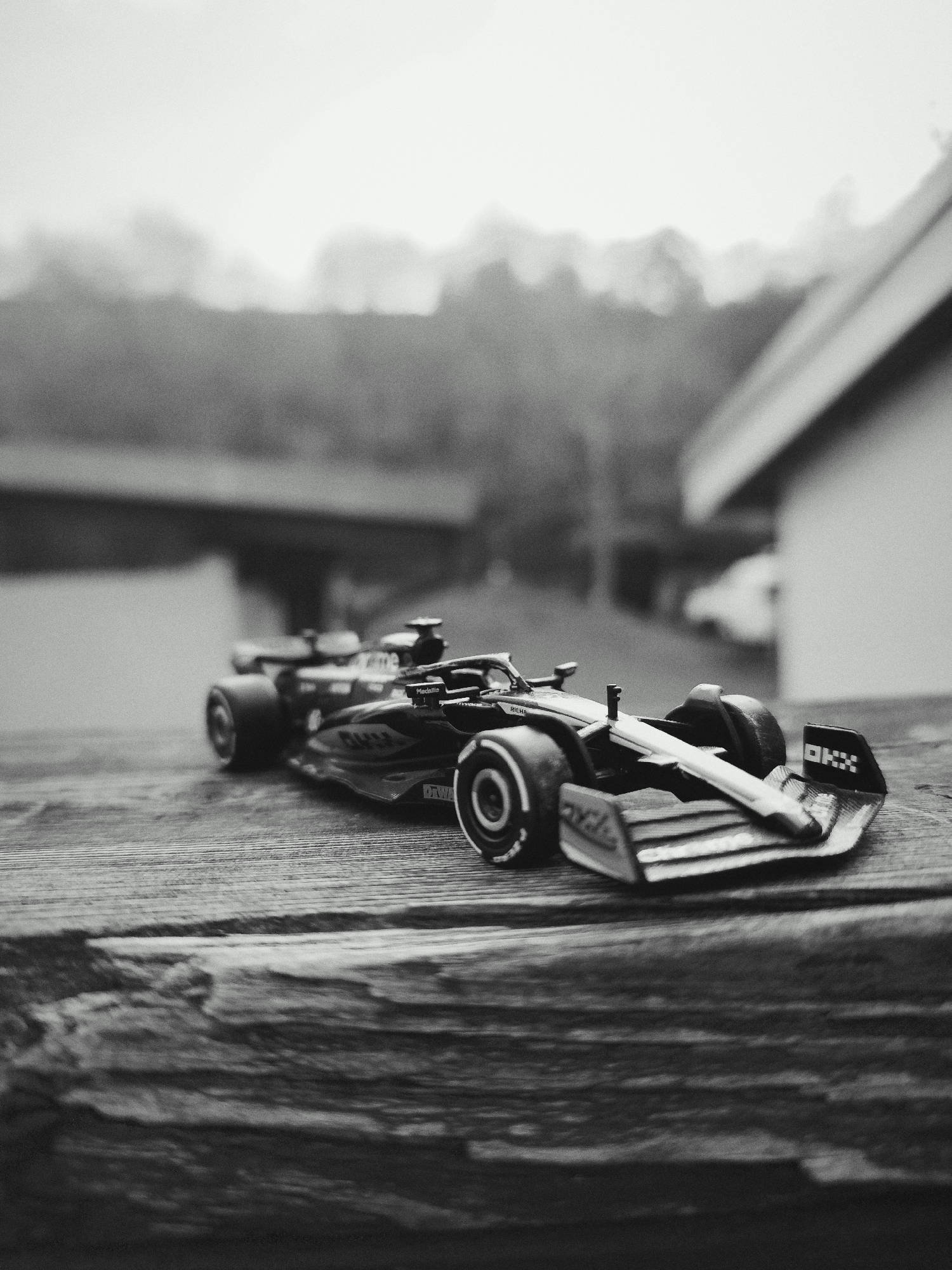 The image displays a black and white, slightly grainy, outdoor scene located in Leidersbach, Germany. The central subject is a dark-colored miniature Formula 1 race car, sharply in focus, positioned diagonally on a weathered wooden surface, likely a deck railing or table. The car model exhibits sponsor decals such as "OKX" on its rear wing, partially visible "RICHARD MILLE" on its sidepod, and "Medallia" on the engine cover, with visible "Pirelli" branding on its tires.
In the blurred midground, to the left and slightly behind the primary car, a second, out-of-focus miniature Formula 1 car is discernible. The background consists of diffused elements: a dark, horizontal structure to the left, and a light-colored building with a visible roofline on the right. Above these structures, a dense line of blurred trees or foliage stretches across the horizon, topped by an overcast sky. No individuals are present, and no actions or interactions are depicted.