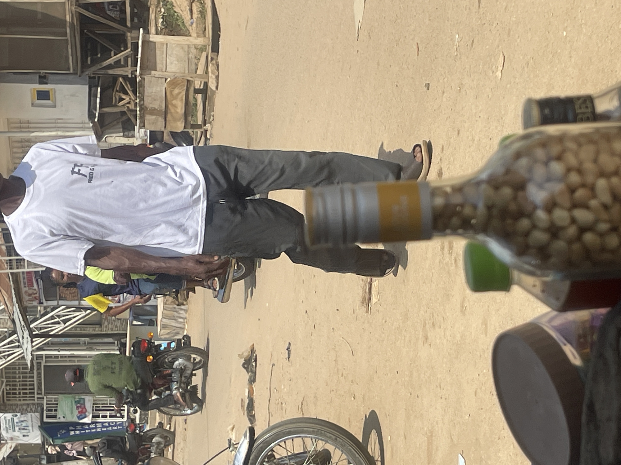 The image depicts a street scene in Abuja, Nigeria, under bright daylight. In the foreground, out of focus, are several bottles, one of which contains a visible quantity of small, light-colored beans or nuts. Behind these, a person is walking away from the camera, wearing a white t-shirt with a graphic logo and dark gray pants. They are also wearing light-colored sandals. Further back, a person in a green jacket on a motorcycle is visible, as is another individual in a yellow and blue vest. The background shows buildings with corrugated metal roofs, some appearing to be shops or market stalls, and a dusty, unpaved road. Several other motorcycles are parked or in motion on the street, suggesting a busy, active environment. The overall impression is that of a typical day in a bustling urban setting.