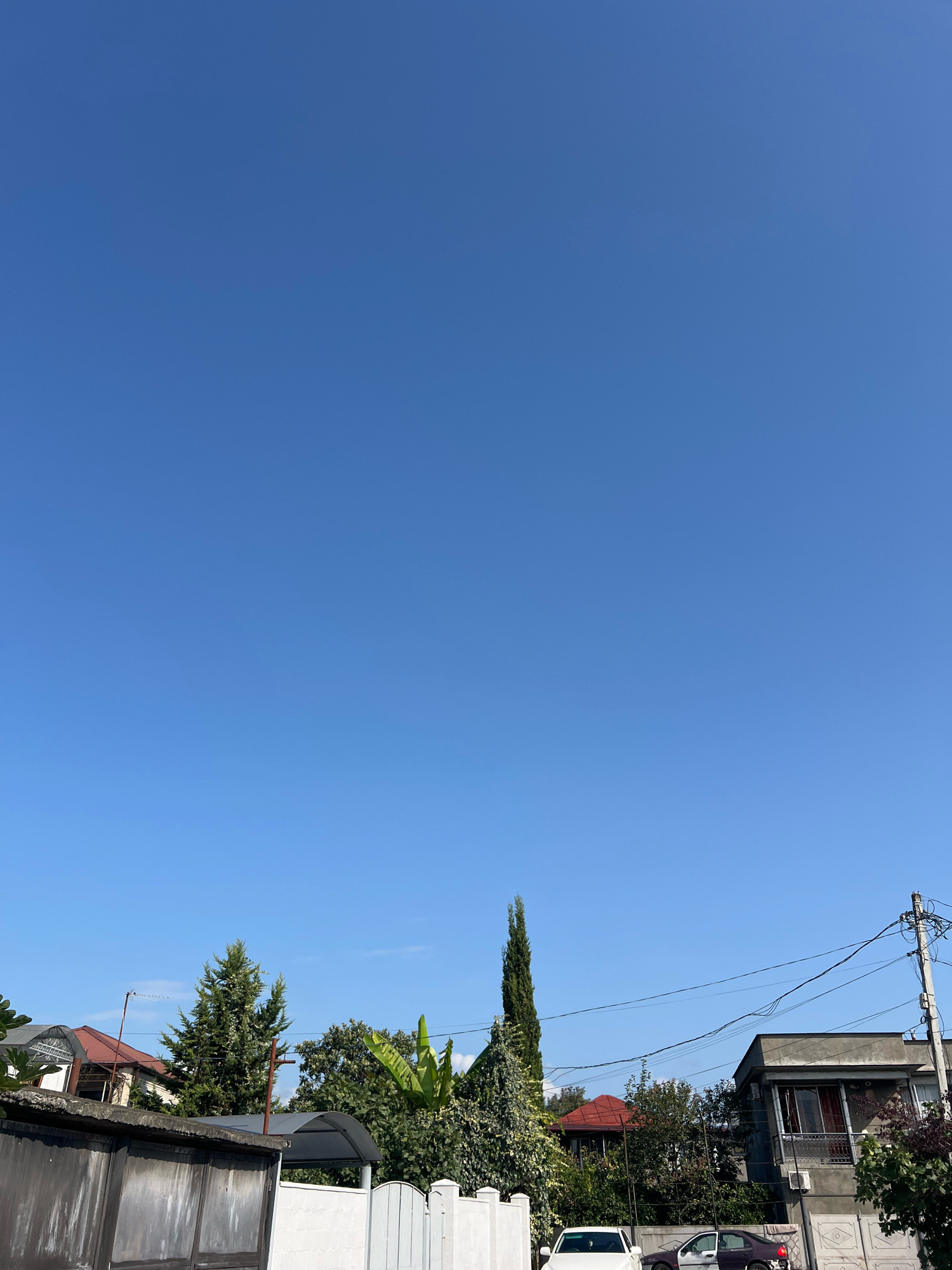 The image captures a clear, bright blue sky over a residential area in Sokhumi, Abkhazia. In the foreground, a section of a grey, weathered wall or fence is visible. Beyond that, a white fence with decorative panels separates properties. Several houses with red and brown roofs are scattered among lush green trees, including a tall, slender cypress tree. A narrow, dark-colored car is parked on the street, with another white car partially visible behind it. Utility poles and wires crisscross the upper portion of the scene, indicating the presence of infrastructure. The overall impression is a tranquil, everyday scene in a semi-urban setting.