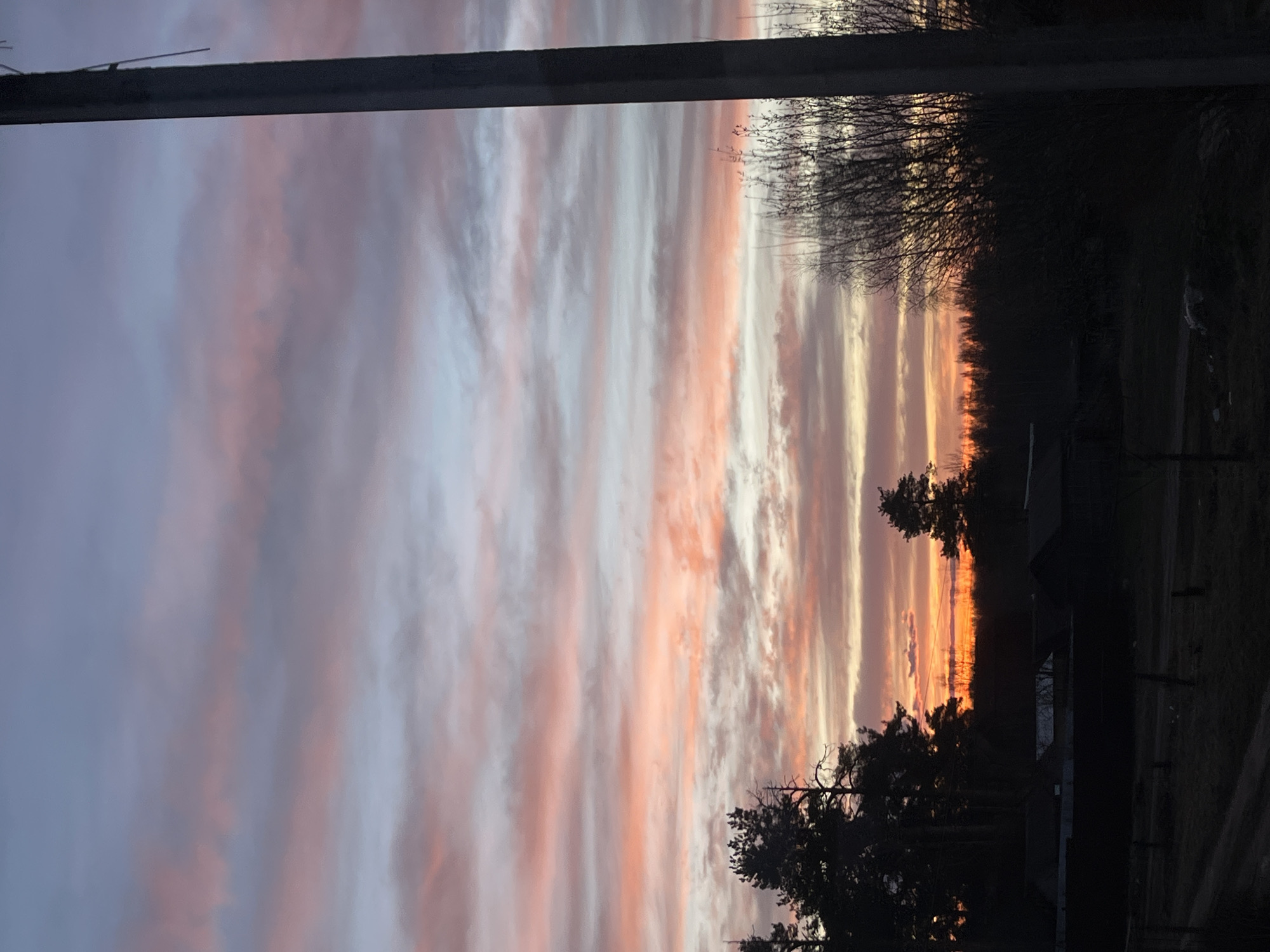 The image depicts a sunset scene viewed from indoors, possibly through a window frame. The sky shows a mix of purple and orange hues in layered clouds. In the foreground, silhouetted trees and roofs of buildings are visible against the sky. The location is Razdolye, Russia, based on the user's request.