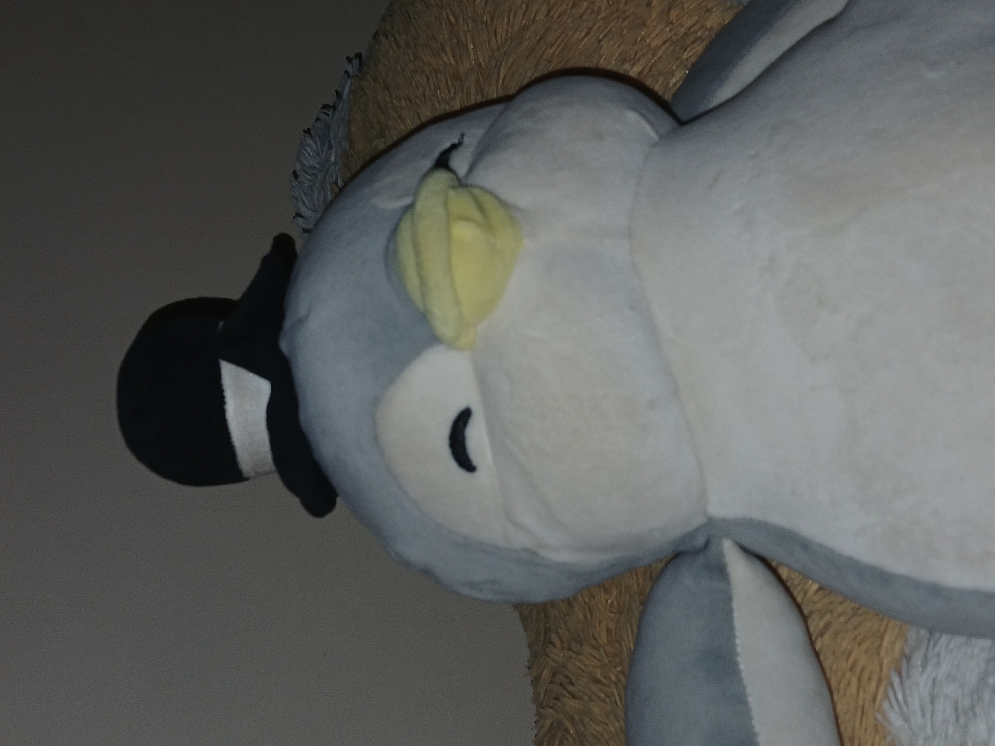 The image displays a large, light grey and white plush toy, possibly resembling an owl or another bird, resting on a textured, fuzzy brown surface. The stuffed animal is characterized by a prominent yellow beak or nose area, with black crescent-shaped eyes. On its head, it wears a small black top hat with a white band. The plushie's body appears soft and rounded, indicative of a typical stuffed toy. The background behind the plush toy is a muted, plain grey wall. The lighting in the scene is dim, suggesting either a low-light environment or possibly evening/nighttime. There are no people present, and the stuffed animal is stationary. No text or specific location cues for Bengaluru, India, are discernible within the image. The scene conveys a calm, indoor setting.