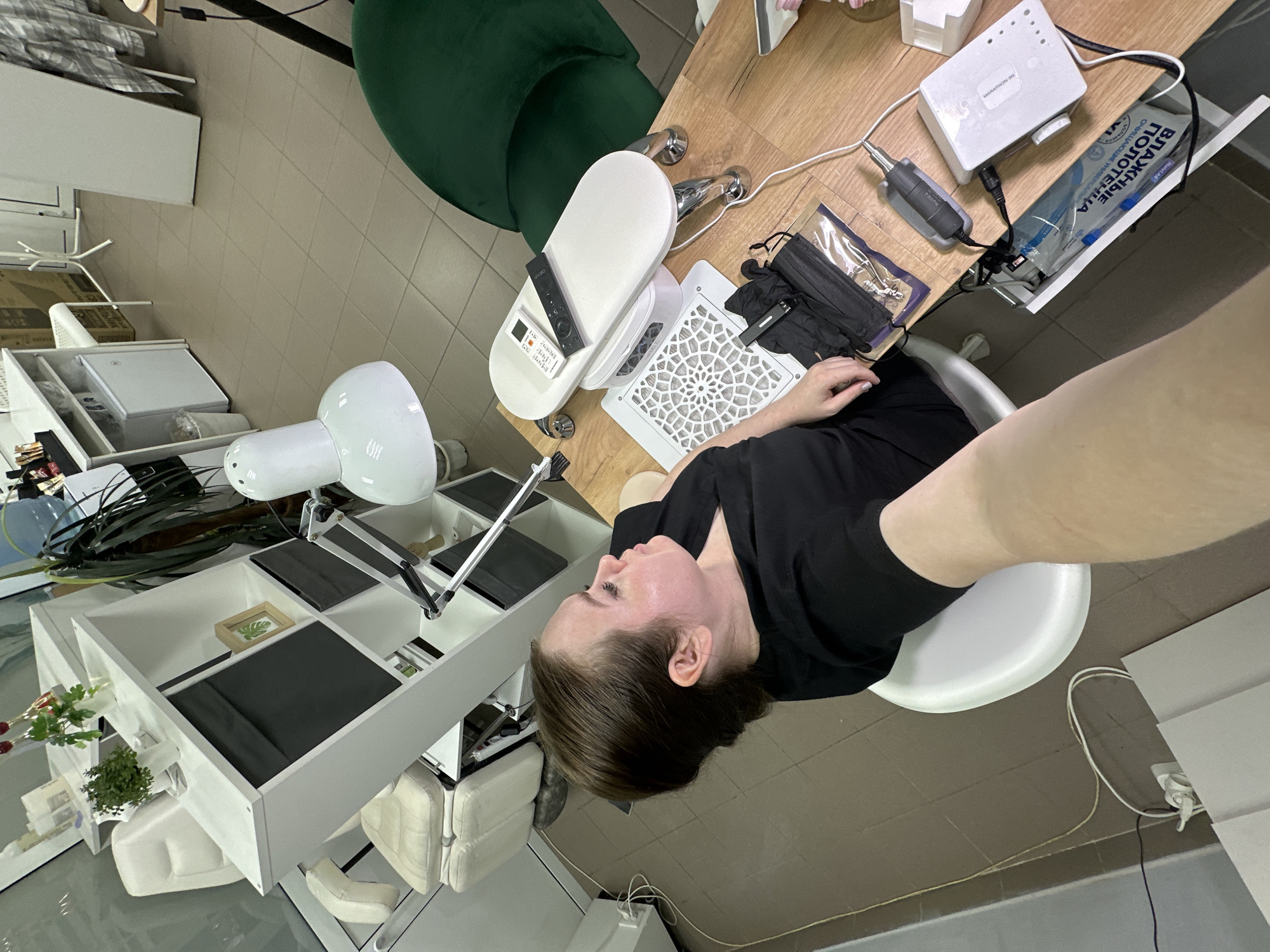 This image depicts an indoor scene within a beauty salon or nail studio, located in the city of Chaikovsky, Russia. A female individual, appearing to be in her 30s-40s, is seated on a white stool in the foreground, facing right in profile. She has light brown hair pulled back, and her complexion shows some redness. She is wearing a black short-sleeved top, and her right forearm extends towards the viewer, suggesting she is capturing a selfie or a close-up photograph of the setting.
The primary focus is a wooden workstation table. On the table, various tools for nail care are visible: a white, oval-shaped armrest with two remote controls (one white, one black), a square white dust collector/fan with an ornate grid, and a white Marathon 3N brand nail drill machine with its handpiece. A clear plastic bag contains black protective gloves, a black surgical face mask, and metal implements (likely cuticle pushers or nippers). A white power strip is also present, connecting the drill machine. A partially opened drawer reveals a packet labeled "ВЛАЖНЫЕ ПОЛОТЕНЦА" (Wet Towels) in Russian. A large white desk lamp with a prominent head is positioned over the workstation.
In the mid-ground to the left, a white modular shelving unit contains dark grey fabric storage boxes, a small potted green plant, a framed leaf print, and two red decorative champagne flutes. Behind this unit, a cream-colored pedicure chair/recliner is partially visible. Further back, a white mini-fridge and a white waste bin are located under a white table. To the right of the workstation, a dark green upholstered chair with a high back is visible. The floor throughout the scene is covered with light brown square tiles. In the far background, a white coat stand and a large white panel leaning against a wall are present.