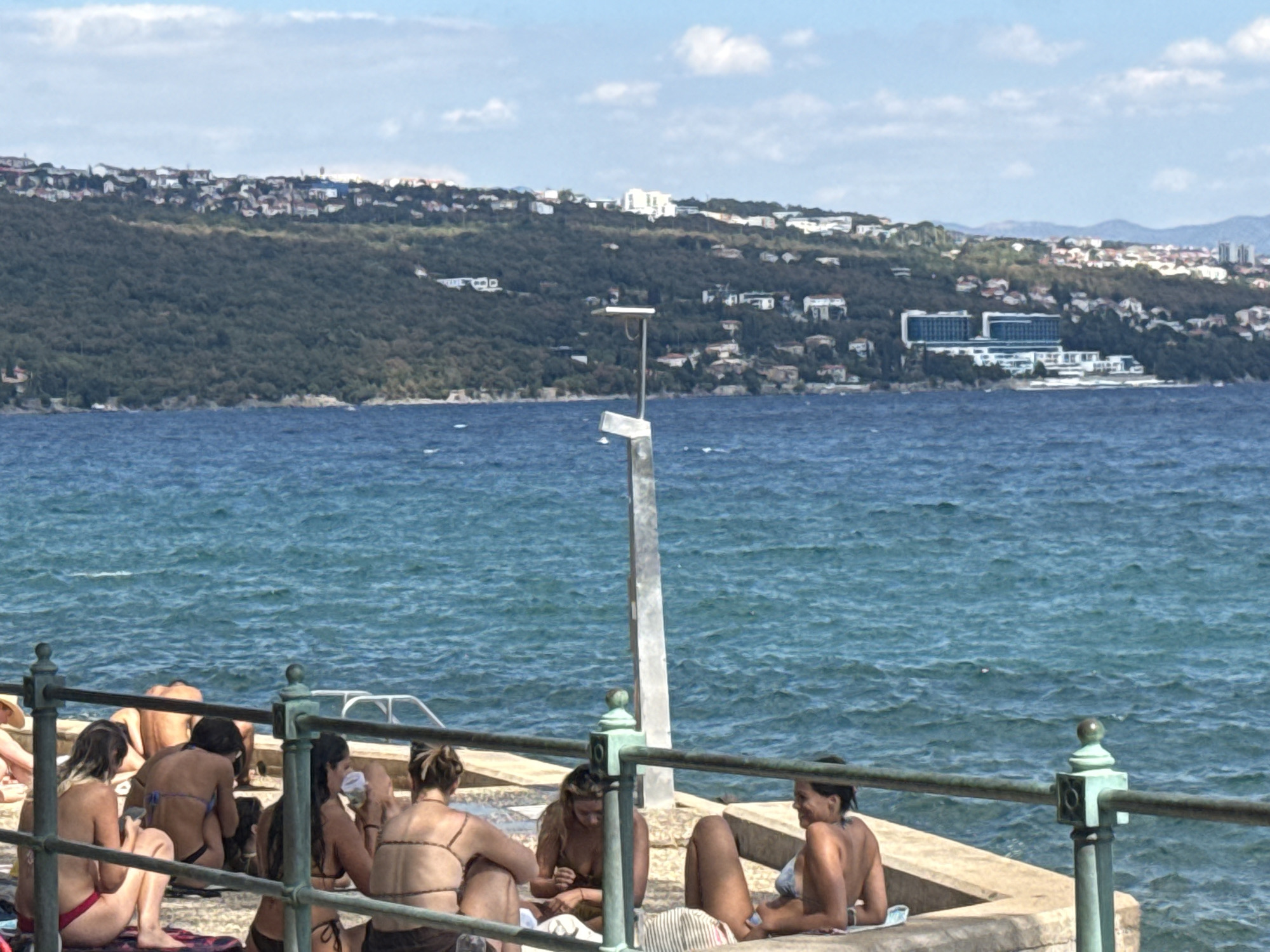 In the city of Opatija, Croatia, a group of people are sunbathing on a concrete pier overlooking the Adriatic Sea. The water is a vibrant blue with small waves. In the background, a lush green hillside is dotted with houses and buildings, with a large white resort hotel visible near the shoreline. The sky is partly cloudy with a bright sun. Several individuals, primarily women, are seated or lying down on towels, wearing swimwear. Some are looking at their phones, while others are engaged in conversation. A metal railing runs along the edge of the pier, with decorative green posts. A tall, metallic pole with a light fixture stands on the pier.