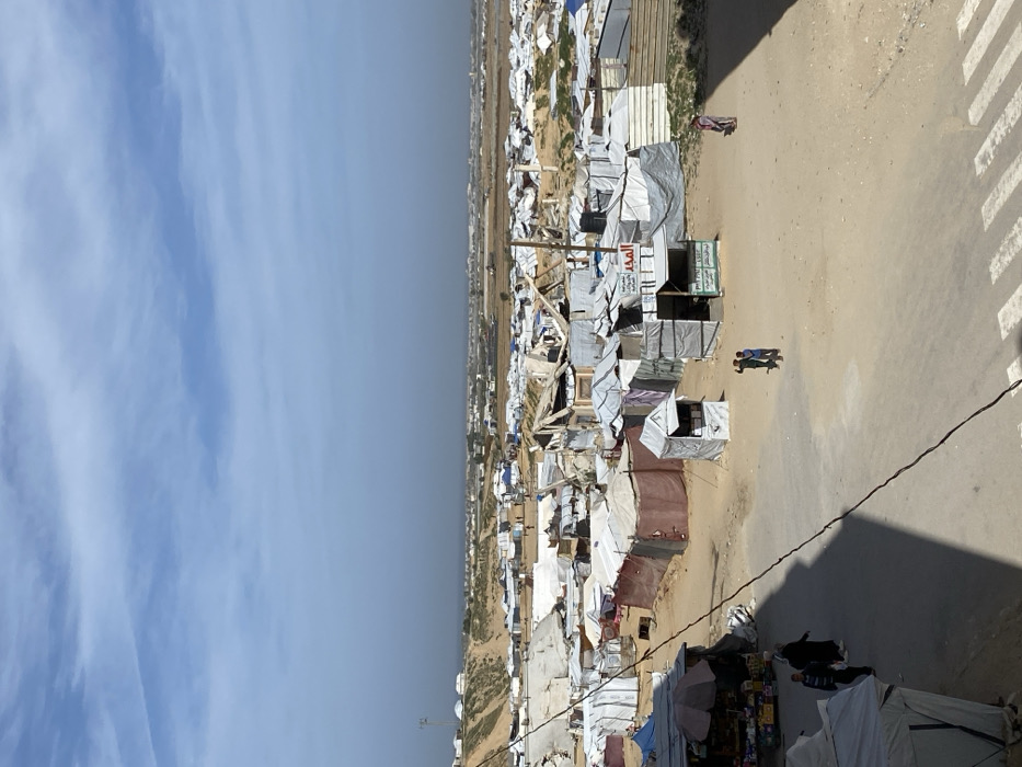 The image captures an elevated view of a sprawling temporary settlement, consistent with a displacement camp, in Khan Yunis, Palestinian Territory, under a partially cloudy blue sky. The foreground features an unpaved sandy road with a visible pedestrian crosswalk in the bottom right and a dark diagonal cable partially obscuring the lower left.
On the road, four individuals are visible: two figures walk left near a market stall topped with a dark umbrella and displaying various goods; two individuals (likely children) stand together in the center; and a lone figure in a patterned garment stands to the right.
The midground is densely packed with makeshift shelters constructed primarily from white, grey, and brown tarpaulins, fabrics, and salvaged materials such as corrugated metal. Many structures show signs of significant damage, with torn coverings, exposed frameworks, and partial collapse. Several small, booth-like structures, serving as kiosks or shops, are visible, some with awnings and Arabic signage, including "المجد" (Al-Majd), "الخير والبركة" (Goodness and Blessing), and "مستلزمات منزلية" (Household supplies). A wooden pole stands upright within this cluster of shelters.
In the background, the settlement extends across undulating, arid terrain into the distance, with more scattered temporary structures and barren land visible towards the horizon. A tall communication mast or cell tower is discernible on a distant hill to the far left.