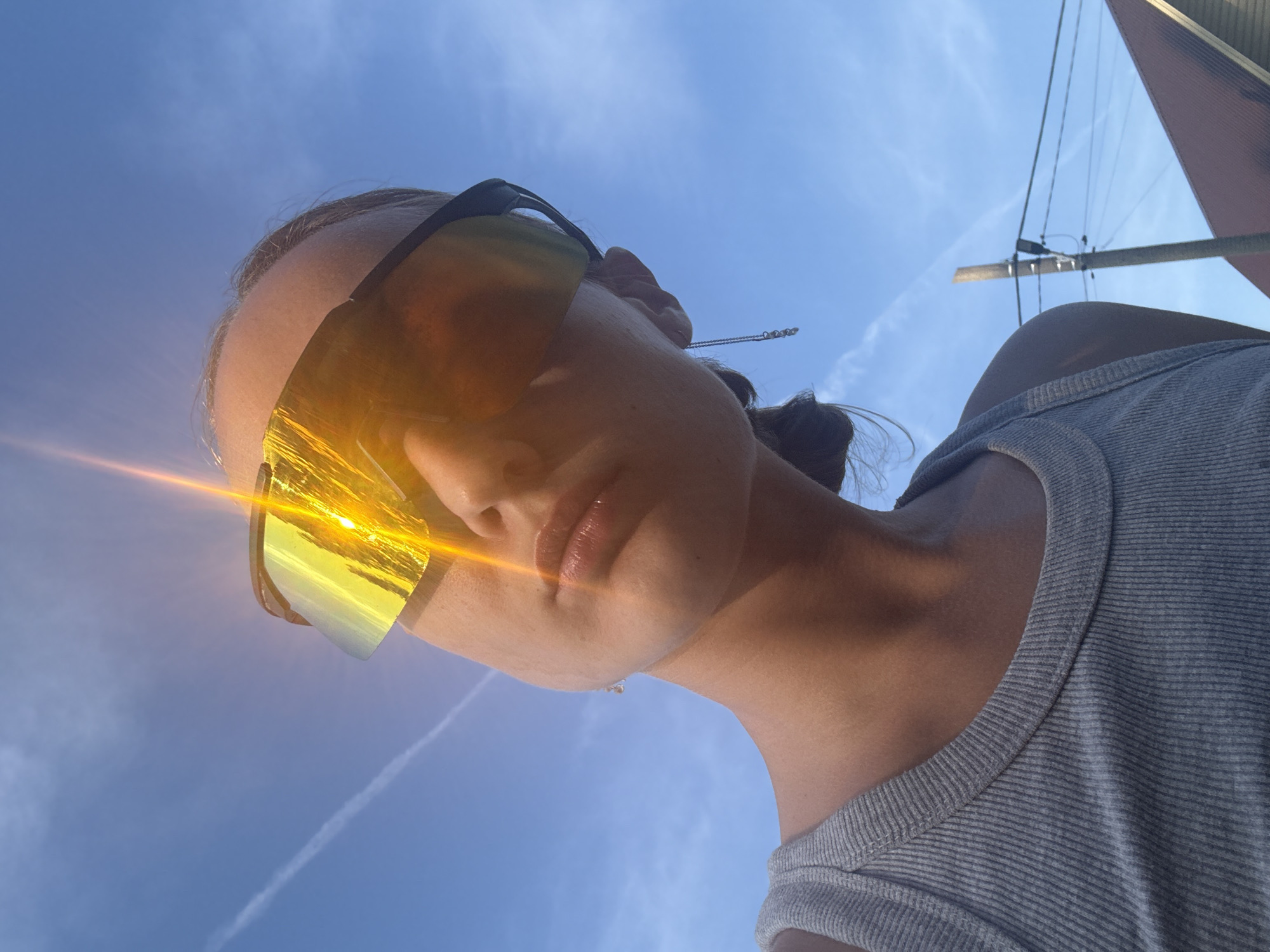 A young woman, seen from a low angle, is looking upwards against a clear blue sky with thin clouds. She is wearing a grey ribbed tank top and large, yellow-tinted visor sunglasses. The sun is prominently reflecting off the sunglasses, creating a bright glare and a lens flare effect. A thin silver earring with small pearls hangs from her ear. The background shows a utility pole with wires and the roof of a building. The overall impression is a casual, outdoor scene, possibly in the city of Podshivalovo, Russia, given the context provided.