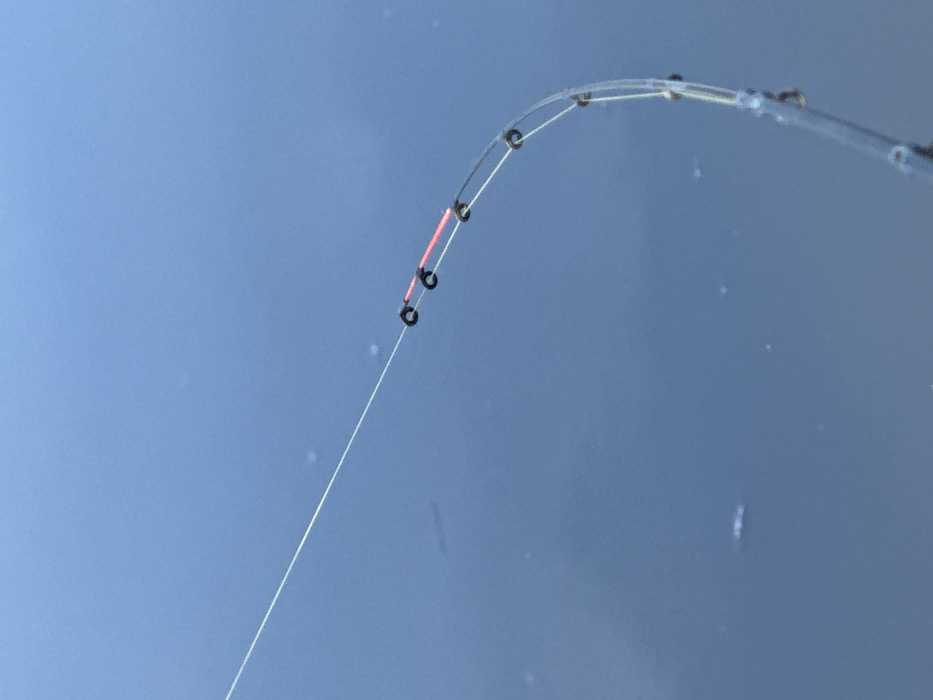 A close-up, overhead view depicts a segment of a fishing line and rod tip positioned over a calm body of water. A transparent monofilament fishing line extends diagonally from the upper left, threading through several small, dark-colored circular line guides. A short, cylindrical, bright orange-red marker or stopper is visible on the line, situated between two of these guides. The line then curves downwards, continuing through additional guides affixed to a translucent, flexible, rod-like structure, likely the tip section of a fishing rod. Small water droplets are present on this translucent rod section. The background consists of a uniform, deep blue-gray water surface, showing minimal disturbance. No individuals or specific landmarks are visible in the frame. The scene is observed in the city of Ям, Russia.