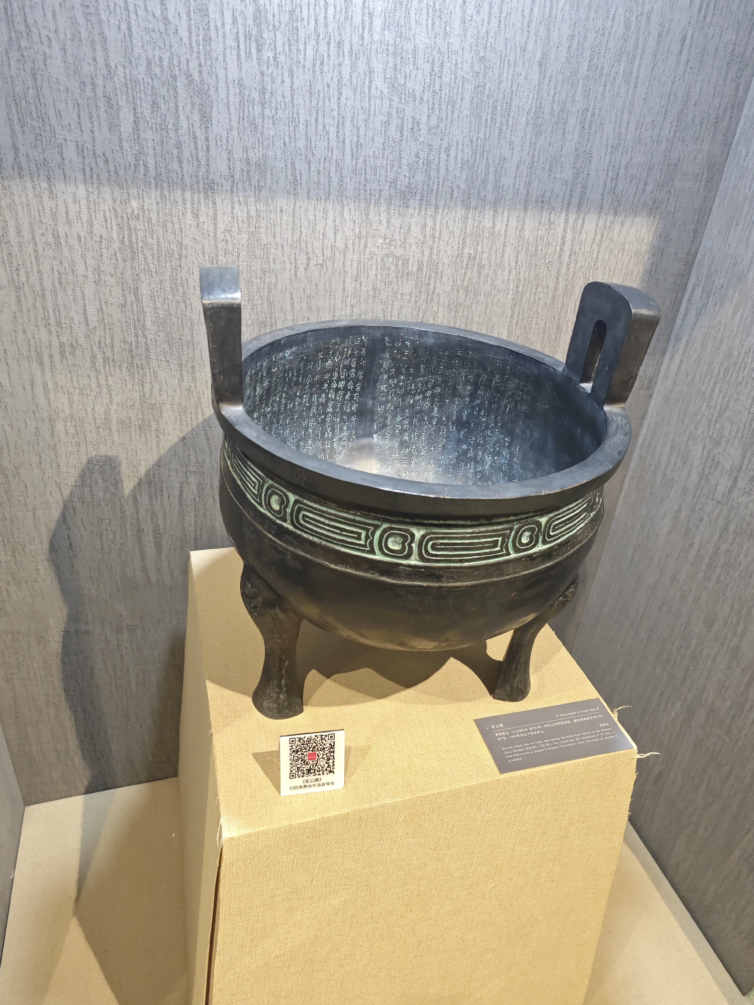 The image shows a bronze artifact displayed in what appears to be a museum setting. The artifact, identified by a nearby label as "Ding-tripod of Duke Mao," is a dark-colored, round vessel with two upright handles and three legs. Inscriptions cover the interior surface of the vessel, and decorative geometric patterns circle its exterior. The artifact rests on a light brown, cloth-covered pedestal. Two labels are visible on the pedestal: one with a QR code and some text in Chinese, and another providing information about the artifact in both English and Chinese. The background features a textured grey wall, suggesting an indoor exhibition space. The specific location, Daxing District in China, is implied but not explicitly seen in the image.