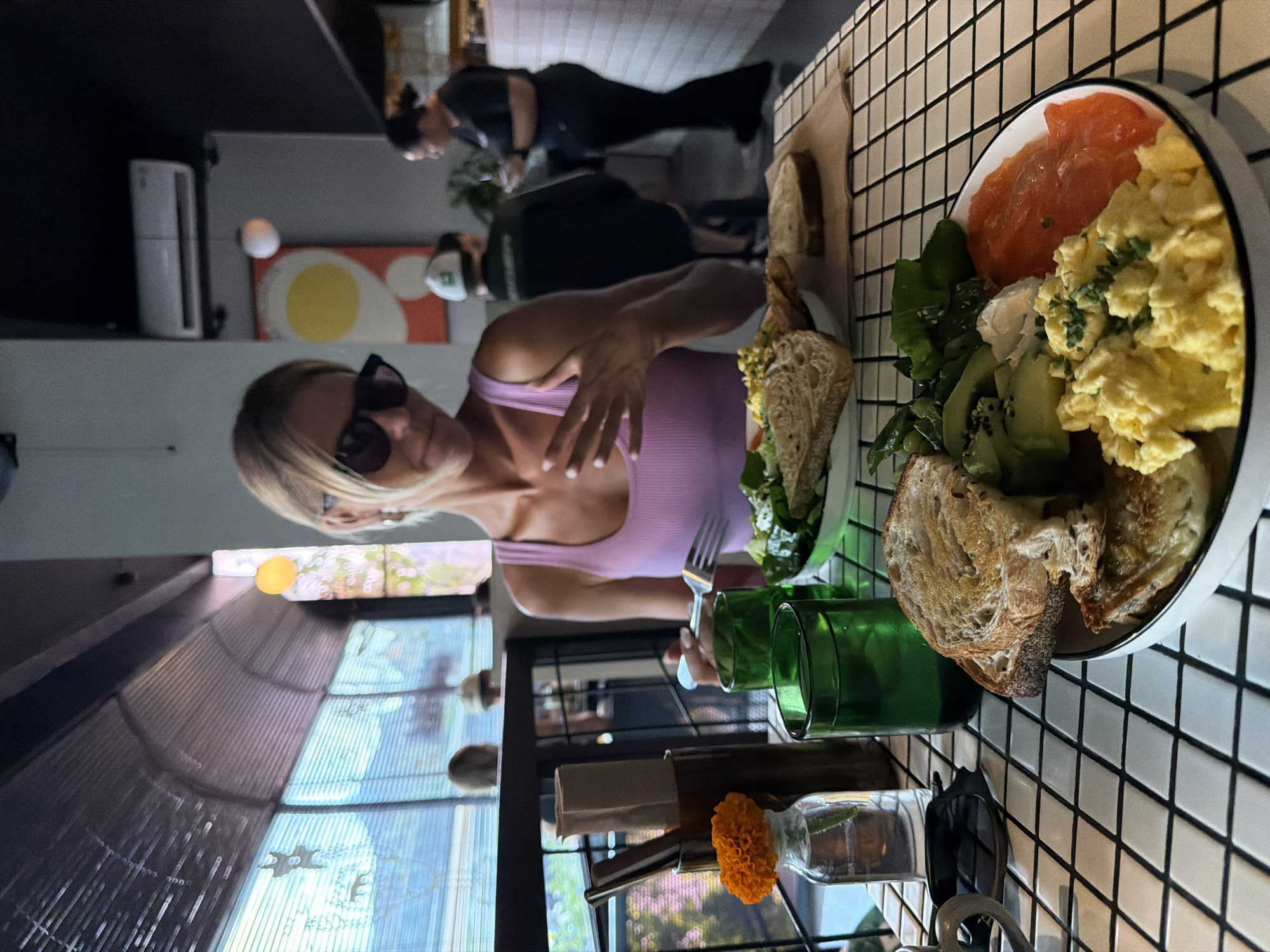 A woman with blonde hair and sunglasses is seated at a table in a restaurant in North Kuta, Indonesia, preparing to eat a meal. In the foreground, a plate holds scrambled eggs, sliced avocado, salmon, and toast. A fork is in the woman's hand. Two green glasses and a vase with an orange flower are on the table. The woman is wearing a pink tank top. In the background, other patrons are visible, and the restaurant features large windows and modern decor.