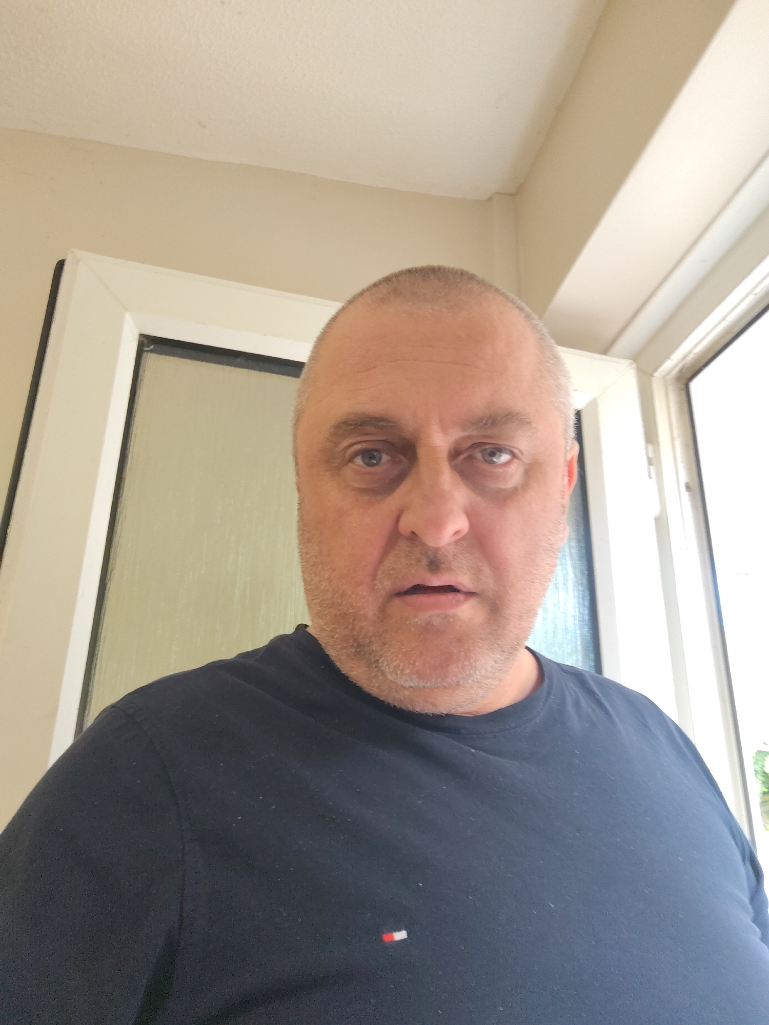 A head-and-shoulders portrait captures a middle-aged man in an indoor setting, likely within Huntingdonshire, United Kingdom. He has closely shaved or bald hair on the top of his head, with short, light-colored stubble covering his jawline and cheeks. The man looks directly at the camera with a neutral to slightly weary expression, his mouth slightly open, and his eyes appearing somewhat heavy-lidded.
He is wearing a dark blue, crew-neck t-shirt. On the left side of his chest, a small, rectangular logo is visible, featuring three horizontal stripes: red, white, and navy, identifying it as the Tommy Hilfiger brand emblem.
The background consists of light-colored, possibly cream or beige, walls and ceiling. To the man's left, a white door frame with a dark inner border partially reveals what appears to be a frosted or obscured glass panel behind it. To his right, another white window or door frame is visible, admitting bright natural light, indicating it is daytime. The overall setting suggests a domestic or residential interior. No specific activity is depicted beyond the man being photographed.