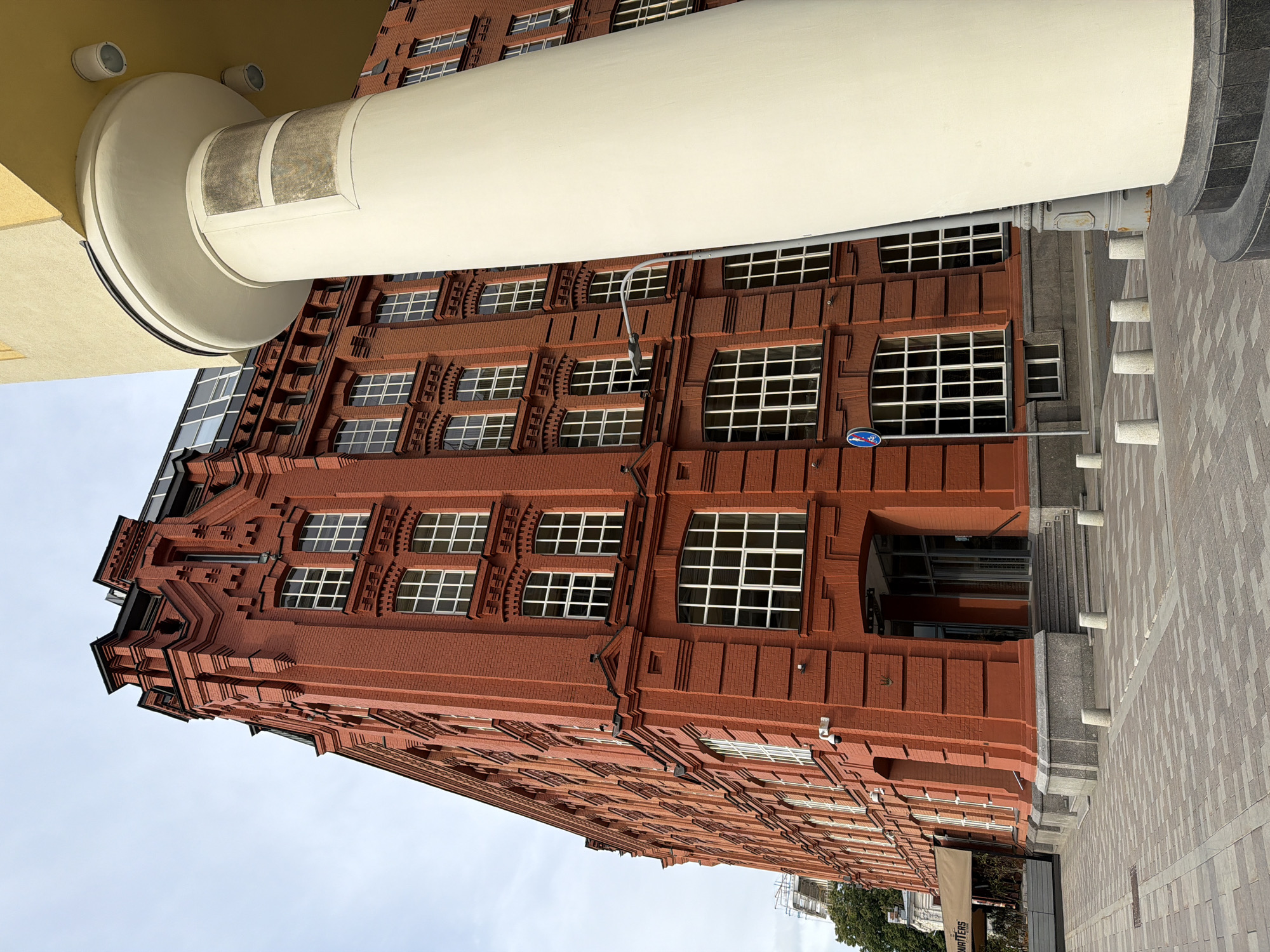 The image depicts a brick building with multiple rows of windows in Moscow, Russia. A large white cylindrical structure runs diagonally across the frame, partially obscuring the building. The building's architectural style features decorative brickwork and arched window frames. The sky is visible in the background. A sidewalk made of light-colored paving stones is on the right side of the image.