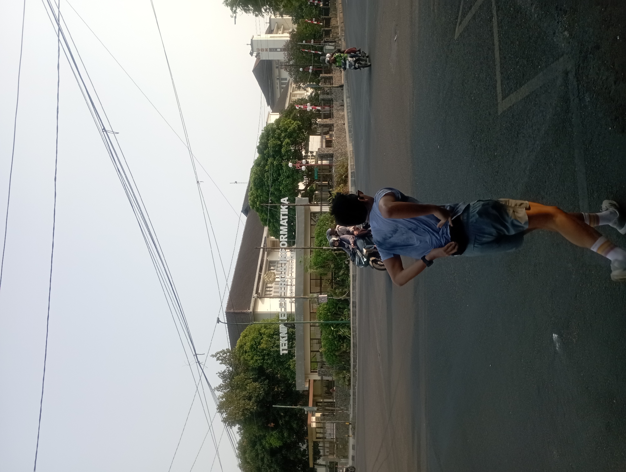 The image captures a street scene in Yogyakarta, Indonesia, at what appears to be late afternoon or early morning due to the low-angled sunlight. In the foreground, a young male wearing a blue t-shirt and grey shorts is seen from behind, with his hands on his lower back, appearing to be stretching or resting during a run. He is wearing white socks with colored stripes and grey athletic shoes. The street is paved and marked with white lines.
In the midground, several motorcycles are present. One motorcycle is in the process of crossing the intersection, carrying two passengers. Another motorcycle is further in the background, also with passengers.
The background features a building complex with signage that reads "TEKNIK ELEKTRO INFORMATIKA". This suggests the location is an educational institution, likely the Faculty of Engineering and Informatics. The building has a light-colored facade and a dark roof. Lush green trees and foliage are visible surrounding the building, and decorative poles with white spherical lights are placed along the sidewalk. The sky is a pale, hazy blue, indicative of either dawn or dusk. Overhead power lines are strung across the frame. The overall impression is an active urban environment with a mix of educational infrastructure and everyday life.