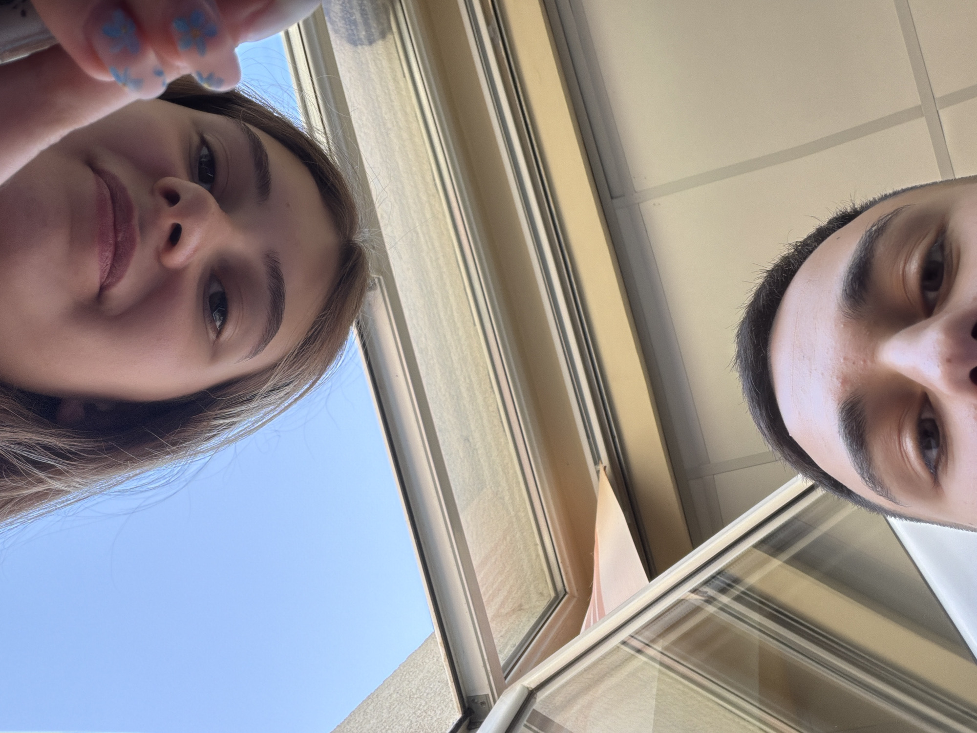 Two people are visible in this low-angle photograph. A woman is on the left, and a man is on the right. The scene is inside, near a window, with a clear blue sky visible outside. Based on the architectural style, this could plausibly be in Panagyurishte, Bulgaria, but further contextual clues would be needed to confirm the location.