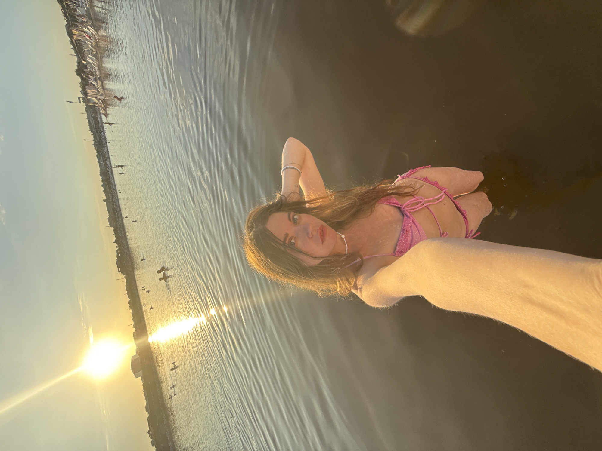 A woman in a pink bikini is taking a selfie in the water at sunset. The sun is setting over a body of water, casting a warm glow on the scene. In the background, there are buildings and people swimming and lounging on the beach. The woman is standing in the water, with her arm extended to take the photo. She is wearing a pink bikini with a white beaded necklace. Her hair is long and brown, and she is looking directly at the camera. The water is calm, with small ripples on the surface. The sky is a clear blue with a few wispy clouds. The overall atmosphere is one of relaxation and enjoyment. The scene is likely the Sol-Iletsk resort, known for its salt lakes and beaches.