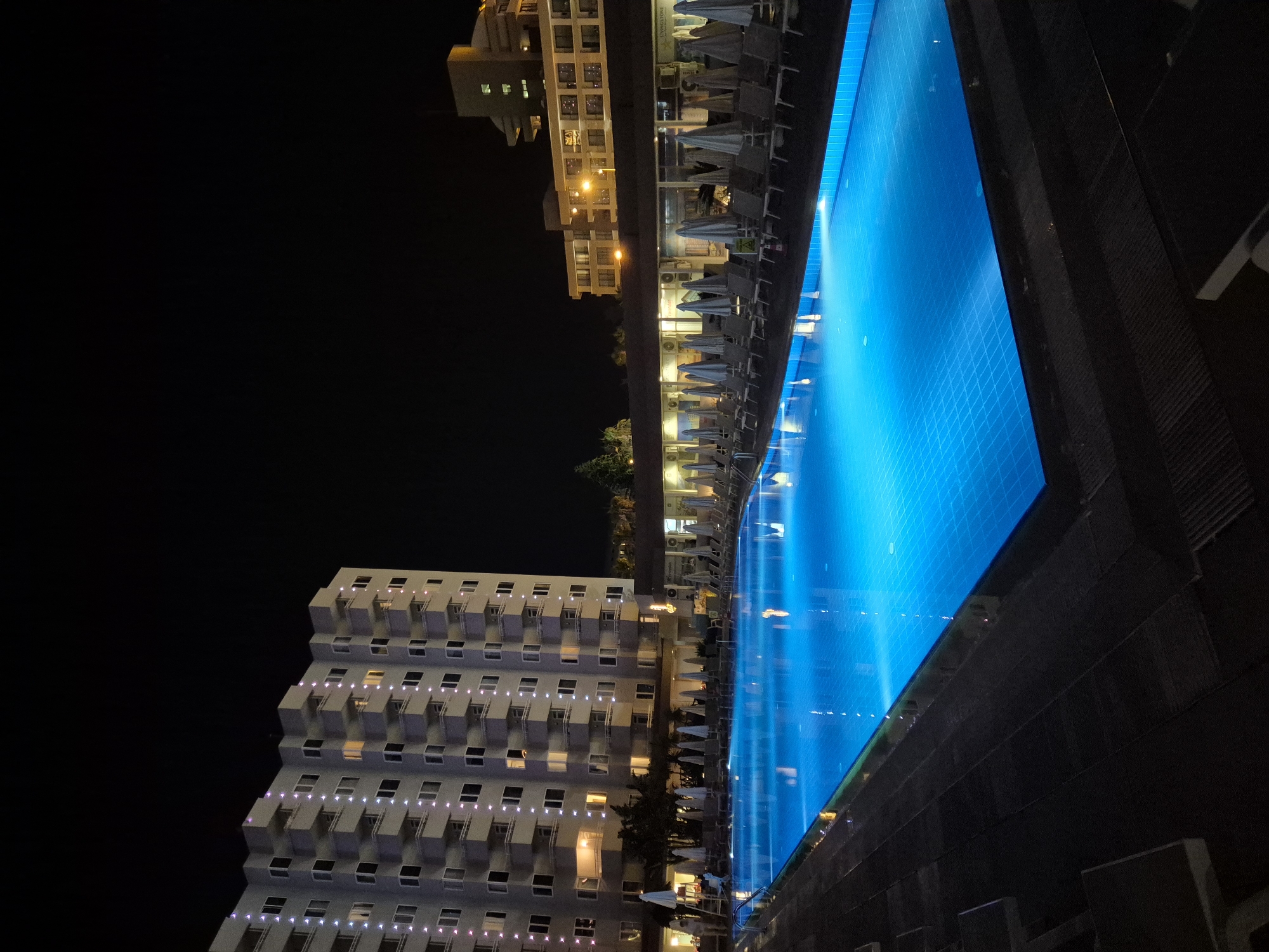 This is a nighttime, eye-level, long shot of a brightly lit swimming pool at a resort. The pool is rectangular and illuminated with blue lights, casting reflections on its surface. Surrounding the pool is a dark deck with numerous lounge chairs and umbrellas, most of which are closed. In the background, a large, multi-story hotel building with a zigzag facade is visible, its windows and balconies outlined with small lights. To the right, another, lower hotel building with illuminated windows and a prominent sign is present. Trees and other foliage are sparsely scattered throughout the scene. The sky is completely dark, indicating it is nighttime.