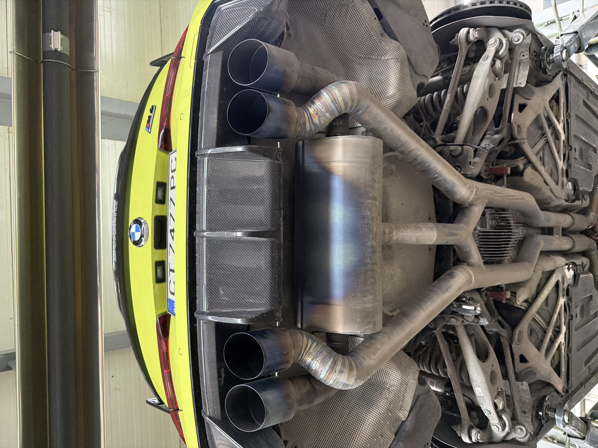 An elevated view captures the underside of a bright yellow/lime green BMW M series car, likely a G80/G82 M4, in what appears to be an automotive workshop. The car is raised on a hoist, with industrial support poles visible on the left side of the frame.
The rear of the vehicle is prominent, showcasing a BMW roundel and a "M PERFORMANCE" sticker on the trunk lid. The license plate reads "CT 7477 PC". A carbon fiber rear diffuser is installed, with four large, dark-finished exhaust tips emerging from it.
The central focus is the aftermarket performance exhaust system. It features intricate, multi-segment piping with visible weld seams, some exhibiting heat discoloration in shades of blue and purple, indicative of titanium construction or heat treatment. A large central muffler also shows similar heat staining. The exhaust pipes route towards the front of the car, with parts of the rear suspension assembly visible, including various control arms and what appears to be a differential. These underbody components appear dusty from use.
The overall scene suggests the car is undergoing maintenance, inspection, or possibly an aftermarket exhaust installation. The lighting is even, typical of an indoor professional garage environment.