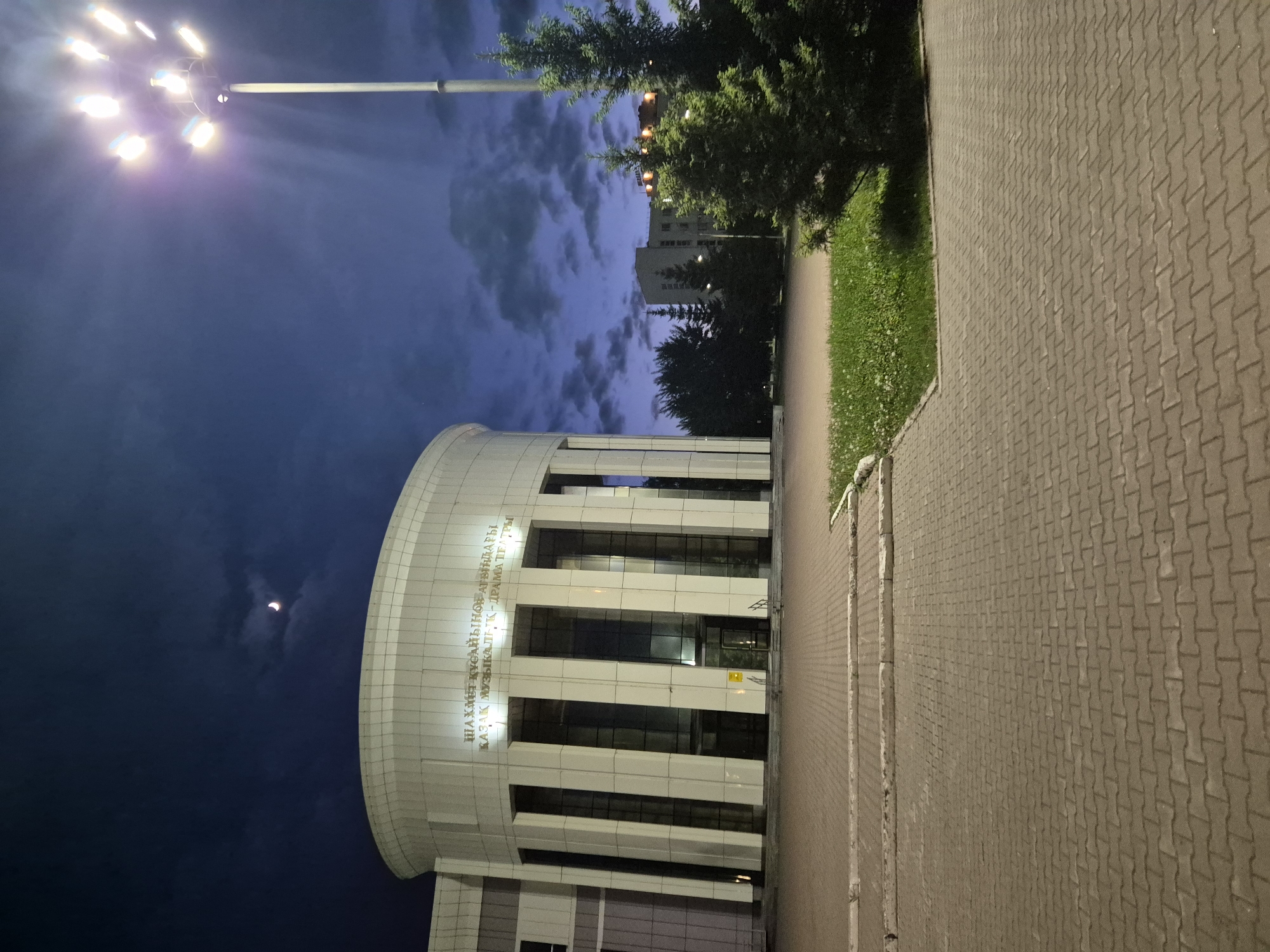 The image depicts the Shakhmet Khusainov Kazakh Drama Theatre in Kokshetau, Kazakhstan, at dusk or night. A multi-bulb streetlight illuminates the area to the left. The theatre's facade is visible, featuring white horizontal stripes and large windows. Text in Kazakh is visible on the curved upper portion of the building. To the right of the theatre is a paved walkway made of interlocking brick pavers, bordered by a strip of grass. The sky shows a gradient of blue and dark clouds, and a partial crescent moon is visible. A tree partially obscures the top of the theatre.