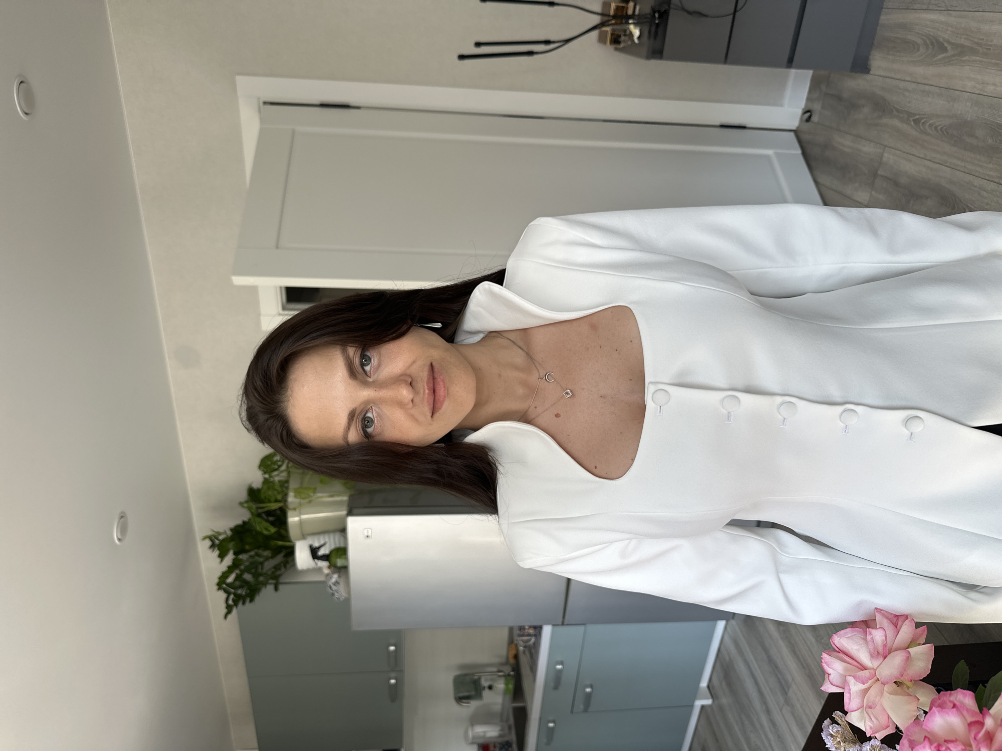 A medium shot captures a woman with long, dark hair standing in a well-lit, contemporary kitchen and living area. She is positioned centrally, facing the camera with a neutral expression. The woman is wearing a white blazer with a distinctive neckline and a row of matching white buttons down the front. She has on a delicate necklace with a crescent moon and a square pendant. In the background, a stainless steel refrigerator is visible to the left, with kitchen cabinets above it. To the right, a portion of a closed white door and a dark dresser with some decorative items are partially visible. The floor appears to be light-colored wood laminate. A bouquet of pink and white flowers is placed in the foreground to the left of the woman. The environment suggests a residential apartment in Murino, Russia.