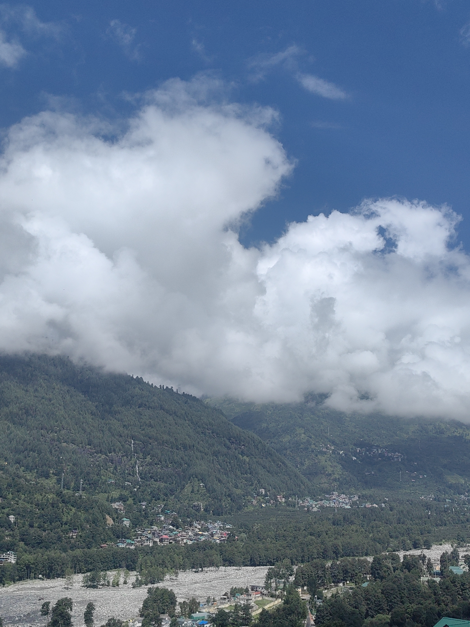 The image depicts a scenic view of Vashisht, India, a town nestled within the Himalayas. The foreground is dominated by a wide, rocky riverbed, with sparse vegetation and trees dotting its expanse. Beyond the riverbed, a dense forest of pine trees covers the lower slopes of a mountain. Further up the mountain, a small town with houses featuring colorful roofs is visible amidst the trees. Several waterfalls cascade down the lush green mountainside. The upper portion of the image is filled with a bright blue sky partially obscured by large, white, fluffy clouds. The overall scene conveys a sense of natural beauty and tranquility.