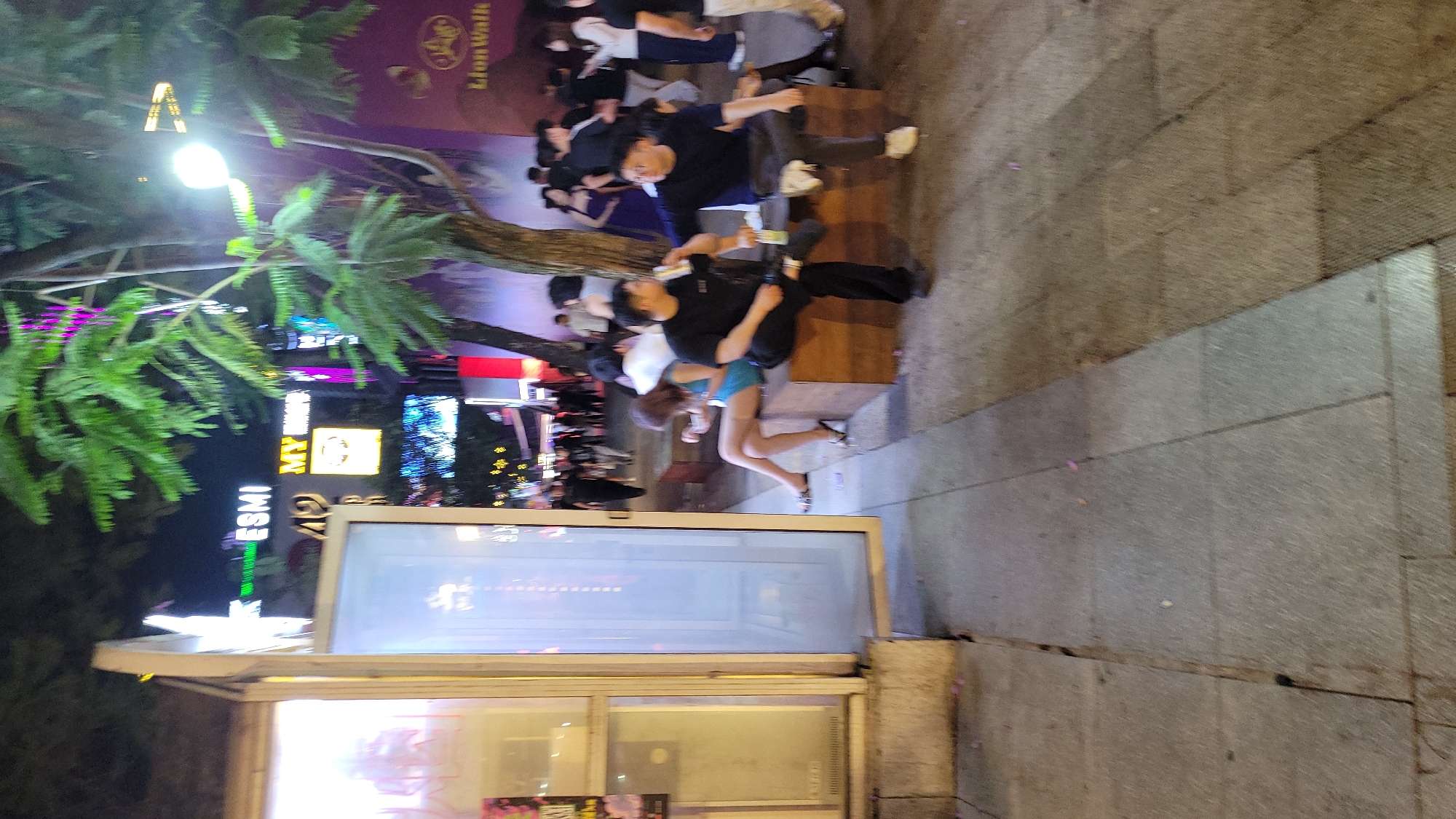 The image depicts a night scene in an urban area, likely a street or entertainment district. In the foreground, there is a structure, possibly a kiosk or small building with a glass window. The window reflects the surrounding environment and some interior elements.
To the left of the structure, there is a street scene with people sitting on benches. Street lights illuminate the area, casting light on the trees and surrounding buildings. There are commercial signs and lighting indicating businesses like "ESMI" and "My Night."
People are sitting and interacting in the background. The street is paved with stone or concrete. The scene appears to be from an urban environment at night.