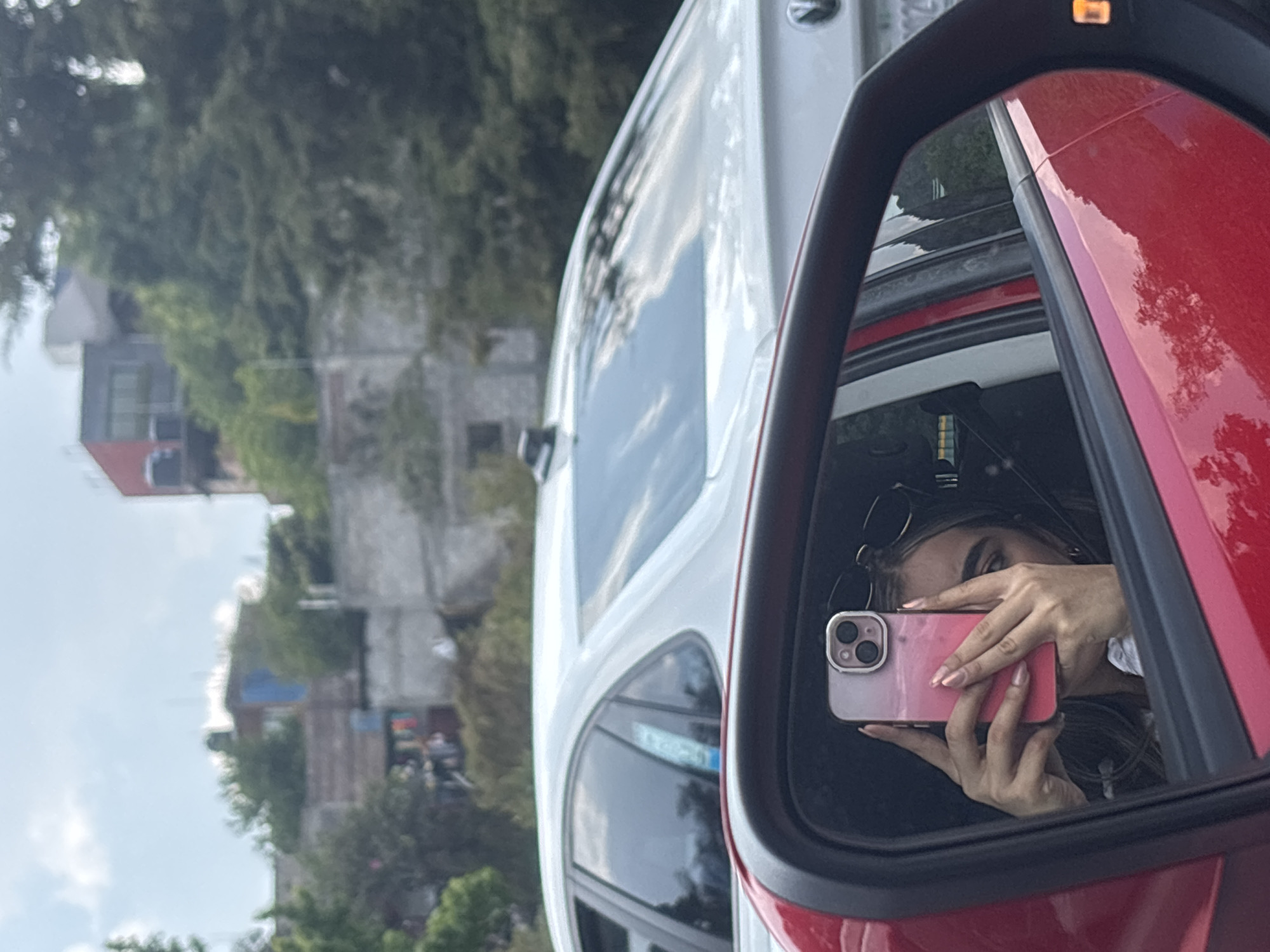 A person takes a photograph of themself in the side-view mirror of a red car. The reflection shows the person holding a pink mobile phone, their face partially visible. The mirror also reflects the interior of the car, including sunglasses resting near the roof. Outside the car, a white vehicle is visible. Further away, a city landscape with buildings and trees can be seen under a cloudy sky. The location is Unknown, Unknown.