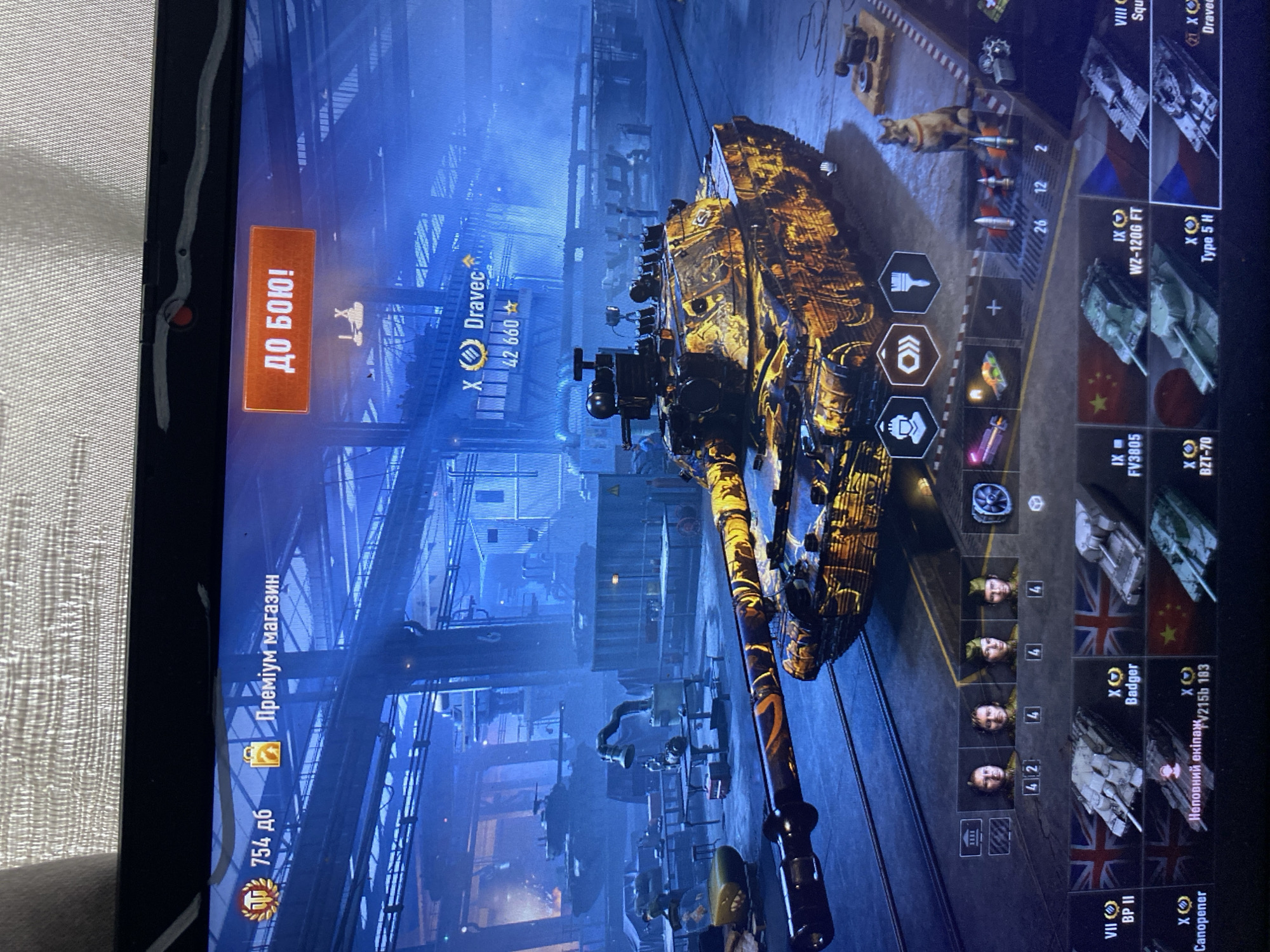 The image shows a screenshot of a video game, likely World of Tanks, displayed on a laptop screen. The game's interface is visible, featuring a tank in the center of the screen with a distinctive orange and black camouflage pattern. In the top left corner of the screen, there's a currency display showing "754 дб" and a button labeled "Преміум магазин". To the right, a prominent orange button reads "ДО БОЮ!", indicating a "To Battle!" function. Below the tank, there's a row of crew portraits and below that, a selection of tanks from various nations, identifiable by their flags and names such as "Badger", "FV3805", "WZ-120G FT", and "BZT-70". A German Shepherd dog sits on the floor to the right of the tank. The background depicts a dimly lit, industrial setting, possibly a hangar or garage with other tanks and machinery. The image is taken from an angle, showing the laptop screen and a textured surface above it, likely a table or desk. There is no indication of the city of Szczecin, Poland, within the image.