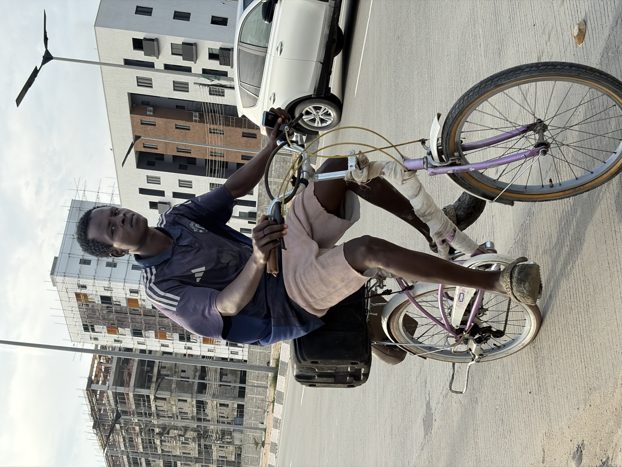 A young Black male is riding a bicycle on a paved road in Alaguntan, Nigeria. He is wearing a dark blue t-shirt with white stripes on the sleeves and light brown shorts. The bicycle has a black box attached to the rear rack and white bandages wrapped around the handlebars and front fork. In the background, a white SUV is driving on the road. Behind the SUV, there are several multi-story buildings under construction, with scaffolding visible. Two modern buildings with windows are also present, one brown and one white. The sky is overcast.