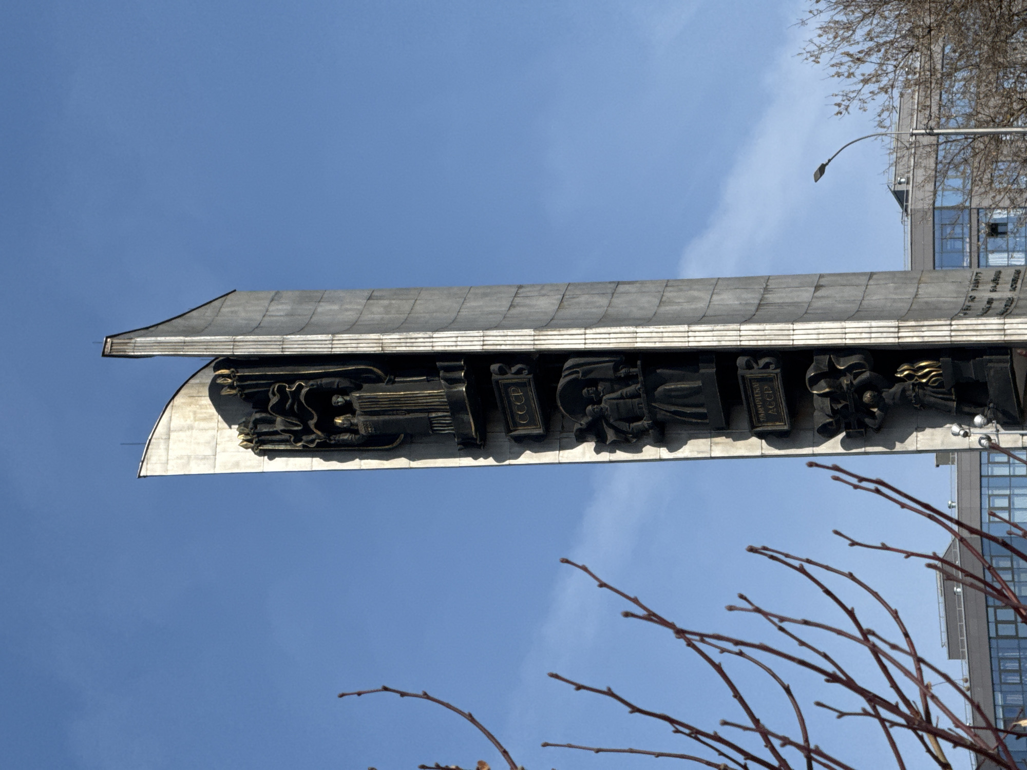 The image shows a monument in Izhevsk, Russia, against a blue sky. The monument has a large concrete overhang with a relief sculpture depicting figures and symbols. The inscription "CCCP" (USSR) is visible on the relief. Nearby are buildings and tree branches. A street lamp is also visible.