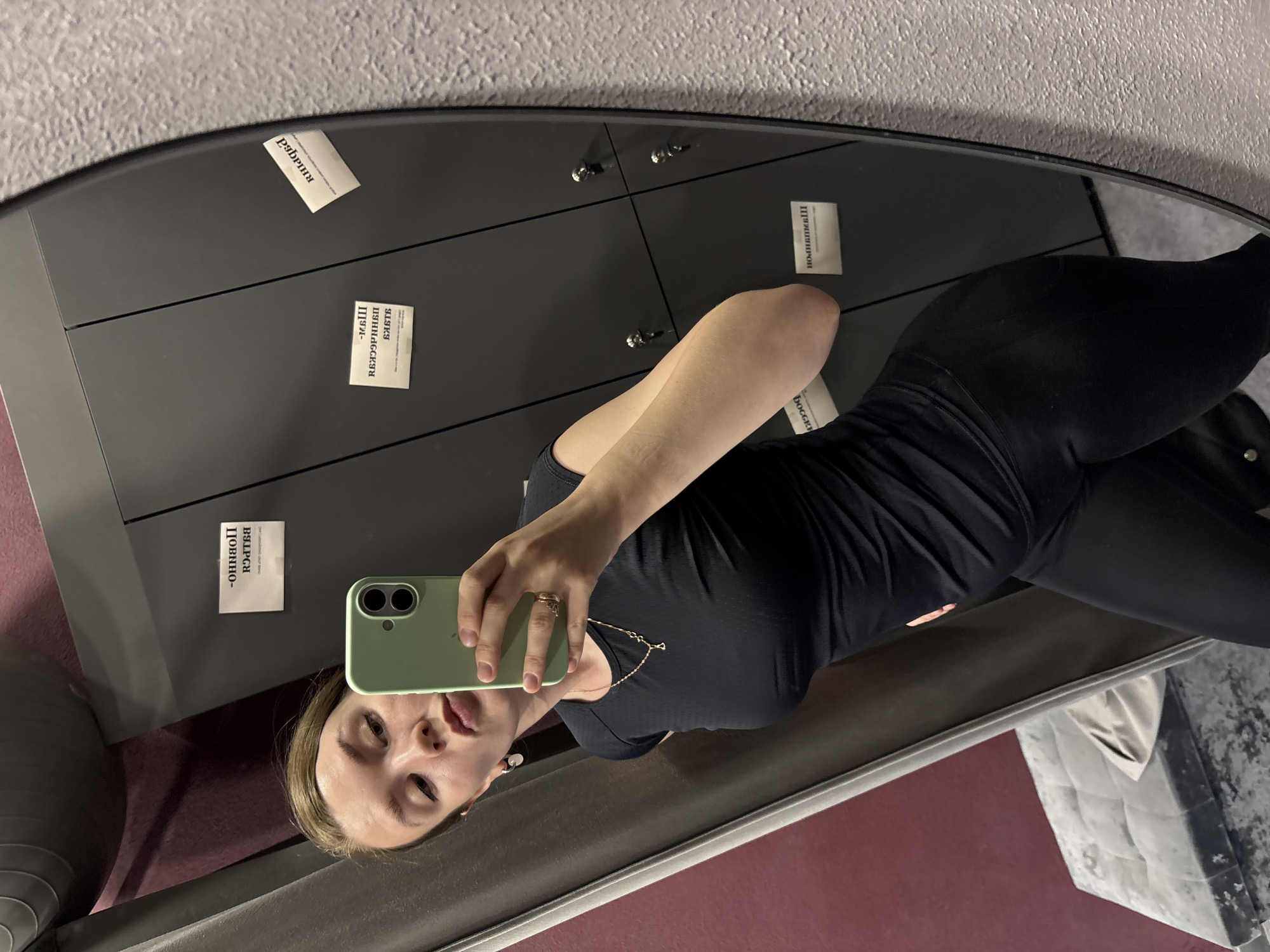 A young woman is captured in a mirror selfie, holding a light green smartphone with her right hand. She is seen from the chest up, with her head slightly tilted and lips slightly parted, looking directly into the camera. She wears a black athletic top and leggings, accessorized with a delicate gold necklace and rings on her right hand.
The setting appears to be an indoor facility, likely a gym or changing room, reflected in the mirror. The background consists of dark gray lockers or storage units, equipped with silver handles or locks. The wall behind the lockers is textured and light gray, while the floor area visible in the reflection is a reddish-brown color.
Several small, white rectangular paper notes are taped onto the lockers. The text on these notes is in Russian, appearing reversed and partially obscured due to the mirror reflection and the image's orientation. Decipherable words and phrases, when mentally corrected for reflection and rotation, include: "РУБРИКА" (Rubric), "ЯВЛЯЙСЯ" (Be/Appear), "ПРИНАДЛЕЖНОСТИ" (Belongings/Accessories), "ПОМНИ" (Remember), "ПРИВЫКАЙ" (Get used to it/Get accustomed), "ОТЛИЧНО" (Excellent/Great), and "ВЫШЕ РЕКОРД" (Higher Record). Partial words like "ШИП." and "ОННО-" are also visible. These messages are consistent with a motivational or informational context found in a fitness establishment in Izhevsk, Russia. The lighting is artificial, characteristic of an indoor environment.