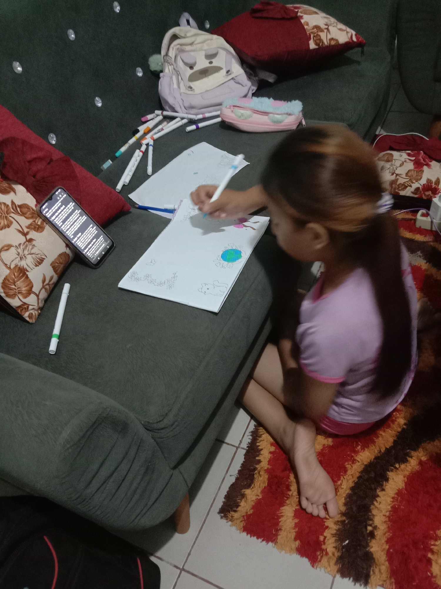 A young girl with long, dark brown hair tied in a ponytail is sitting on a rug next to a dark green sofa in a room. She is wearing a light purple t-shirt and pink shorts. The girl is drawing on a white piece of paper with markers, holding a white marker in her right hand. Next to her on the sofa are several markers, a cell phone, and a white and green marker. On the sofa behind her is a light purple backpack with a bear face design, a pink and blue pencil case, and several pillows. The room appears to be a living area, with a tiled floor and a colorful rug. The setting is likely a home in Kwandang, Indonesia, given the provided location.