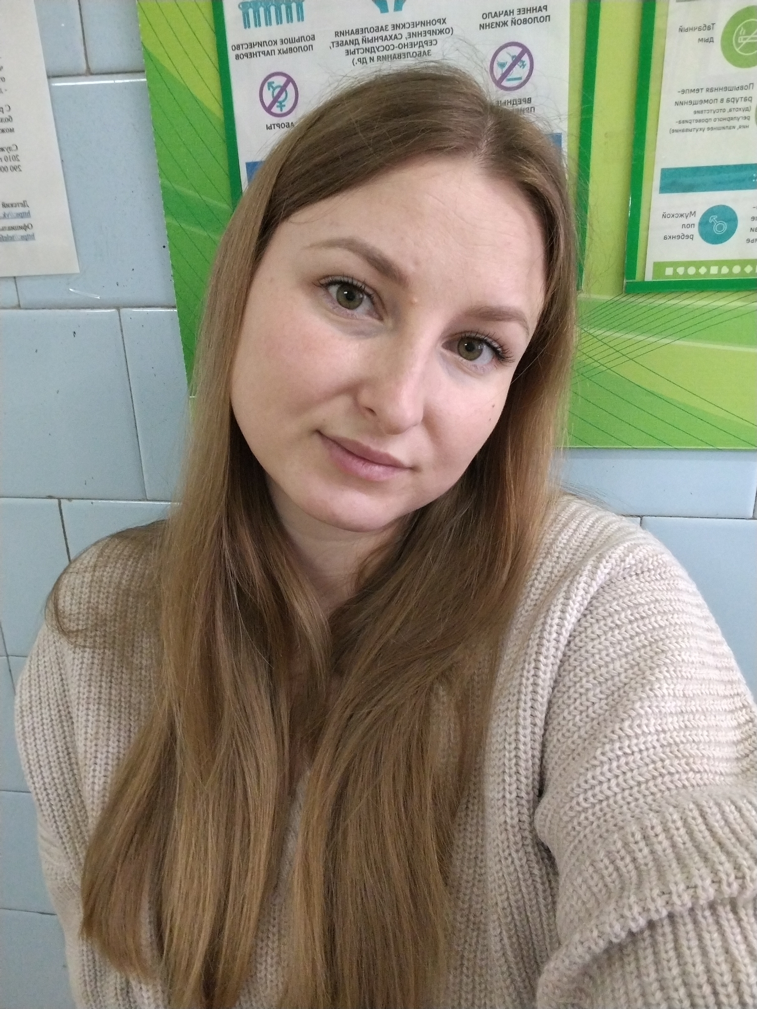 A young woman with long, light brown hair is posing for a selfie indoors. She is wearing a beige knit sweater. The background consists of light blue tiled walls and green and white signs with Russian text and symbols. The environment suggests a clinic or healthcare setting in Chaikovsky, Russia, based on the language and style of the signs.