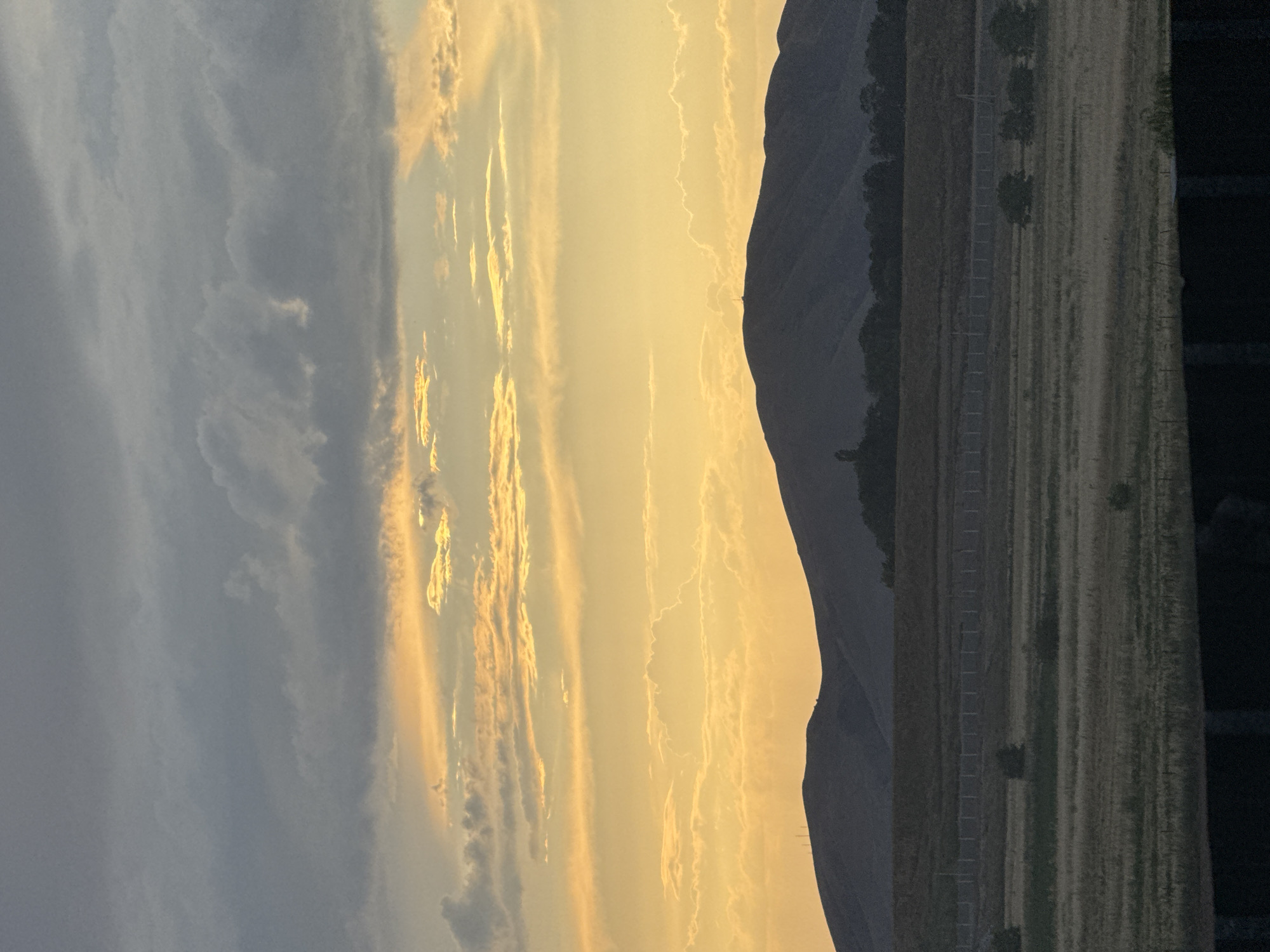 A landscape photograph captures a sunset over rolling hills and a dry, grassy plain. The sky is filled with dramatic clouds, ranging from dark gray at the top to bright orange and yellow hues near the horizon, suggesting the sun is low. The hills in the background are silhouetted against the vibrant sky, with a few small structures, possibly antennas or towers, visible on their peaks. In the foreground, a fence line runs horizontally across the image, separating the field from what appears to be a dark, out-of-focus area at the bottom. The vegetation on the plain is sparse and appears to be dry grass and scattered shrubs. The overall scene evokes a sense of quiet and natural beauty.