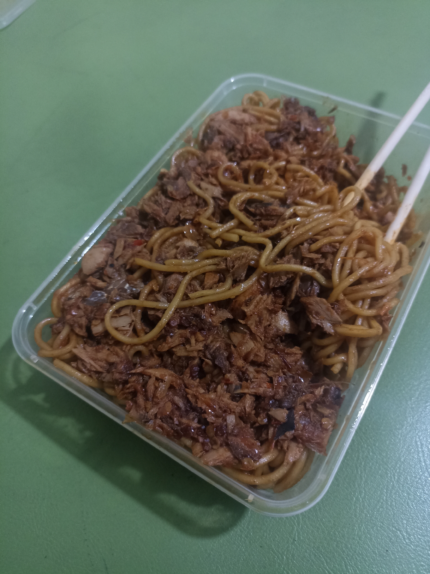 A container of noodles with shredded meat is placed on a green surface. Chopsticks are resting on the container, holding a portion of the noodles. The scene is devoid of any individuals or discernible urban elements.