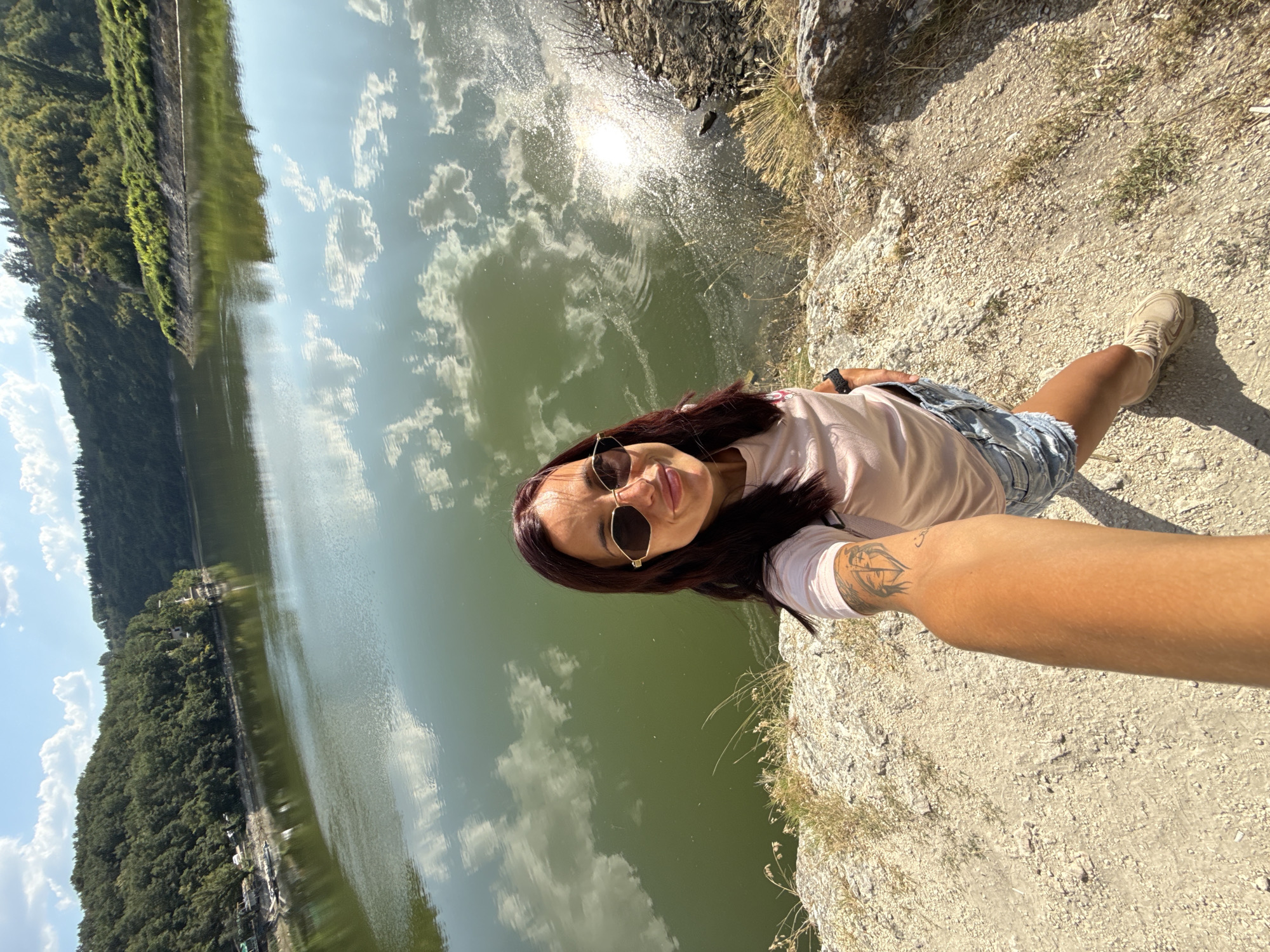 A woman with dark hair and sunglasses is taking a selfie on a rocky embankment overlooking a large body of water. The water reflects the sky and clouds. Trees and hills surround the water. The woman is wearing a pink t-shirt, denim shorts, and light-colored shoes. She has a tattoo on her left arm. The scene appears to be outdoors on a sunny day. This location is likely the dam of the Ivaylovgrad Reservoir, near Pleven, Bulgaria.