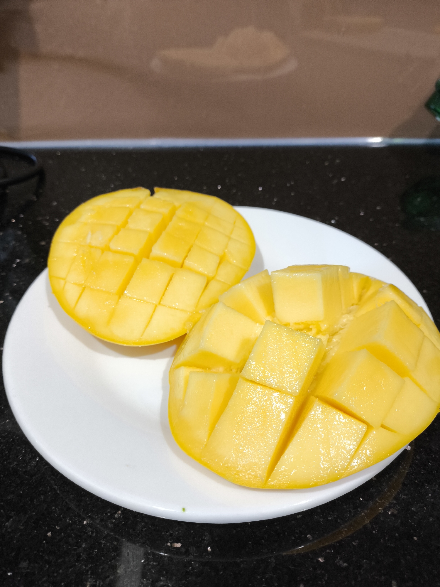 A close-up shot displays two halves of a bright yellow mango, expertly cut into cubes and arranged on a white circular plate. The mangoes are glistening, indicating freshness and ripeness. They are placed on a dark, speckled countertop, possibly granite, which reflects some light. In the background, a tiled backsplash with horizontal lines is visible, in muted brown tones. The overall lighting suggests an indoor setting, possibly a kitchen, and the image appears to be taken during the day. There are no people visible in the frame, and the focus is solely on the prepared fruit. The setting is simple and domestic, with no explicit text present in the image. The arrangement of the mangoes suggests they are ready to be eaten, implying a moment of enjoyment or preparation for a meal.