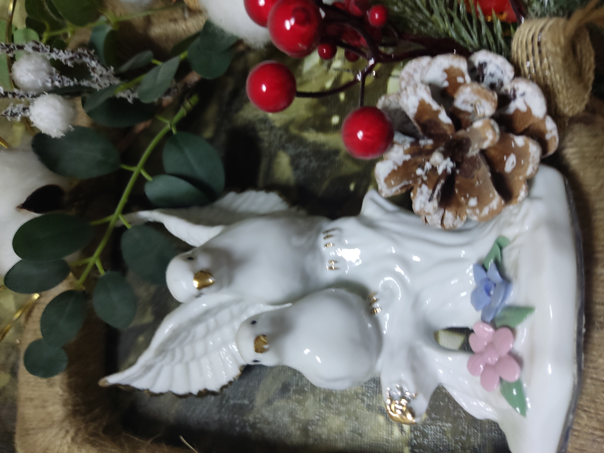 The image shows a close-up of a decorative figurine of two white birds perched on a white base with gold accents. The larger bird has its wings spread, while the smaller bird is perched in front of it. The base is adorned with small pink and blue flowers and green leaves. In the background, there are elements of a nature-themed arrangement, including eucalyptus leaves, red berries, a pinecone dusted with white, and what appears to be cotton or similar fluffy white decorative elements. The overall aesthetic suggests a festive or decorative piece, possibly for a holiday season. The background has a textured, muted pattern. No people are present in the image. The location of this scene in Noginsk, Russia, cannot be determined from the visual information provided.