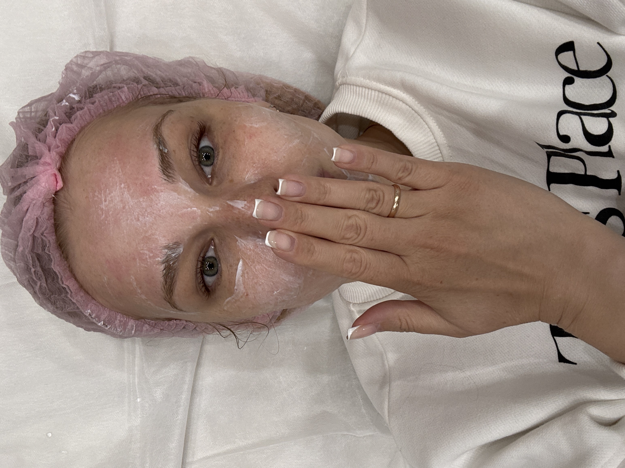 A woman wearing a pink disposable hairnet is lying on a white surface, possibly a treatment table. Her face has a white cream applied to it. Her right hand, adorned with a gold ring, is covering part of her mouth. She's wearing a white sweatshirt with black lettering on the sleeve. The sweatshirt displays the word "Place" in dark letters. The scene is likely indoors, perhaps in a spa or aesthetic clinic. The location in the city of Rusanovo, Russia is not discernible from the image.