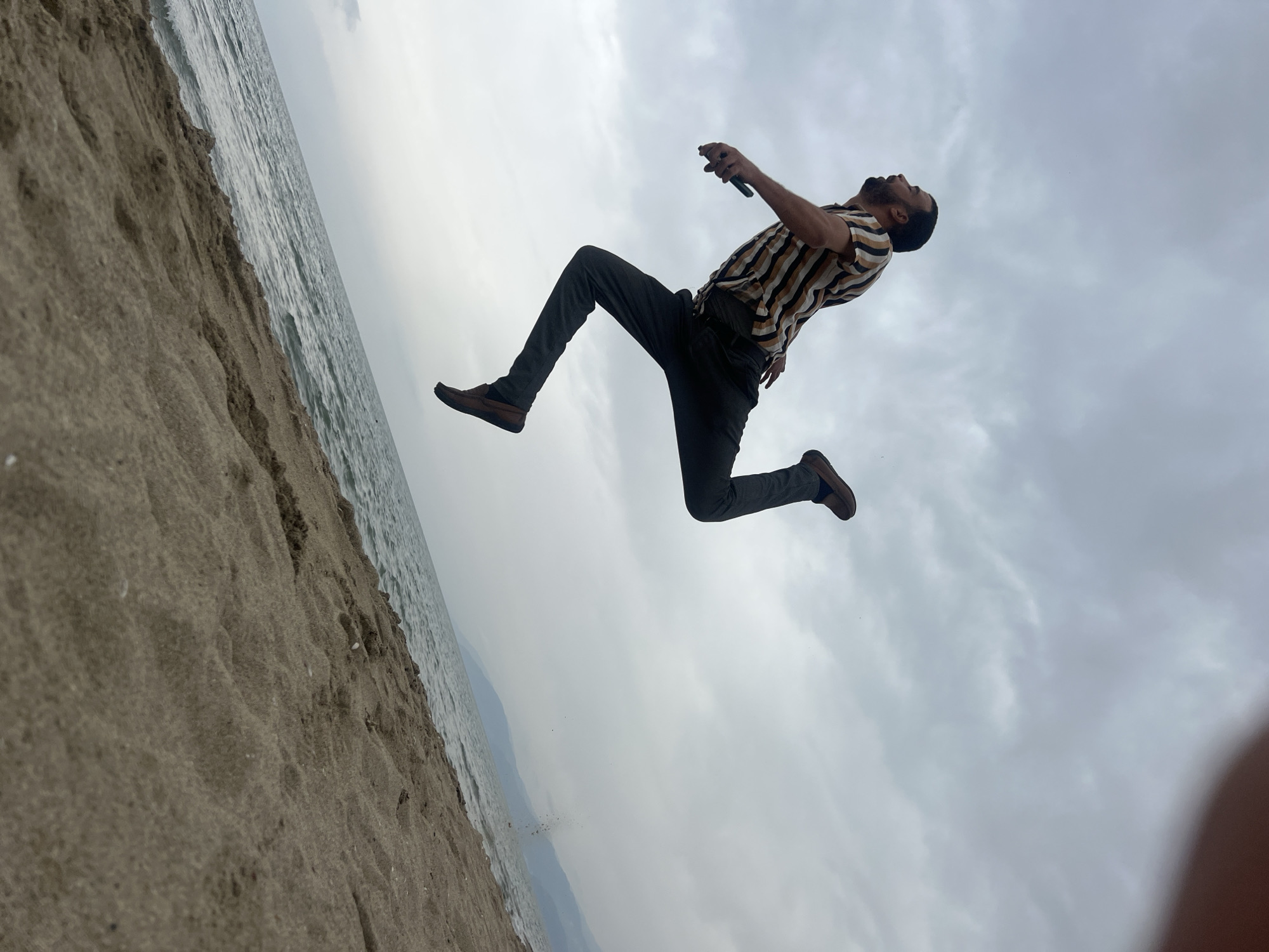 A man is captured mid-air in a dynamic, low-angle shot against an overcast sky at a beach in Martil, Morocco. He is positioned diagonally across the frame, facing upwards and to the right, appearing to leap or float above the ground. He wears a striped short-sleeved collared shirt (light brown, dark brown, and cream stripes), dark trousers, and dark brown shoes. In his right hand, he holds what appears to be a mobile phone, and his head is tilted back, looking up.
Below the man, the scene shows a sandy beach transitioning into the calm waters of the sea. The sand is textured, suggesting footprints or natural undulations. The water appears slightly ruffled, and a faint horizon separates it from the expansive, cloudy sky that fills most of the background. The lighting is soft and diffused due to the overcast conditions, indicating daytime. The image conveys a sense of exhilaration and movement against a natural, coastal backdrop. There is no visible text.