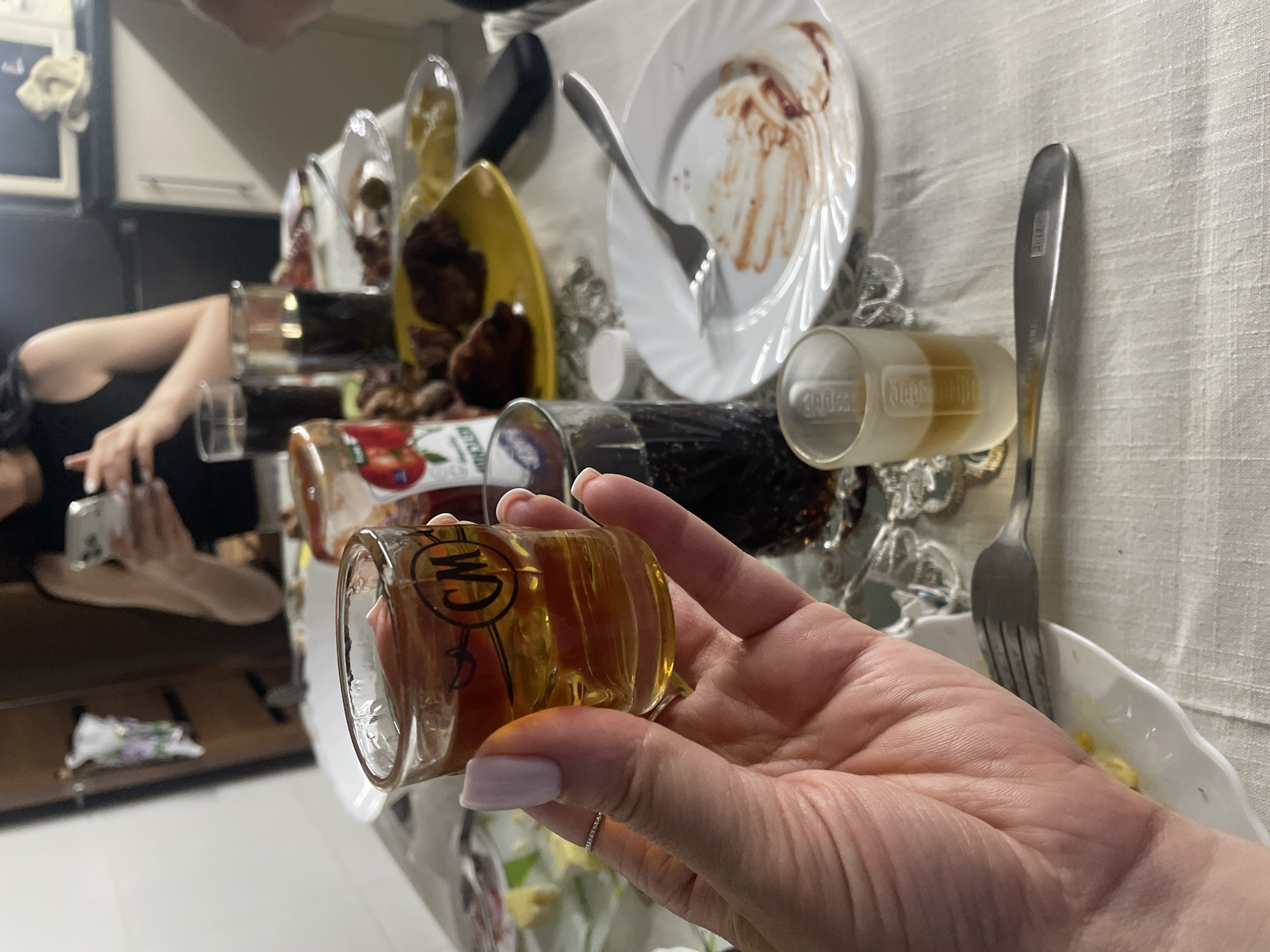The image depicts a close-up of a hand holding a shot glass filled with a amber liquid. The shot glass has a circular emblem with the letters "CM" inside. In the background, a table is set for a meal, with plates, cutlery, glasses of dark liquid, and food items, including what appears to be meat and a jar of ketchup. A person's torso and arms are visible in the upper background, holding a smartphone. The environment suggests an indoor setting, possibly a dining area. No specific landmarks or details indicating the city of Rozsoshentsi, Ukraine, are present in the image.