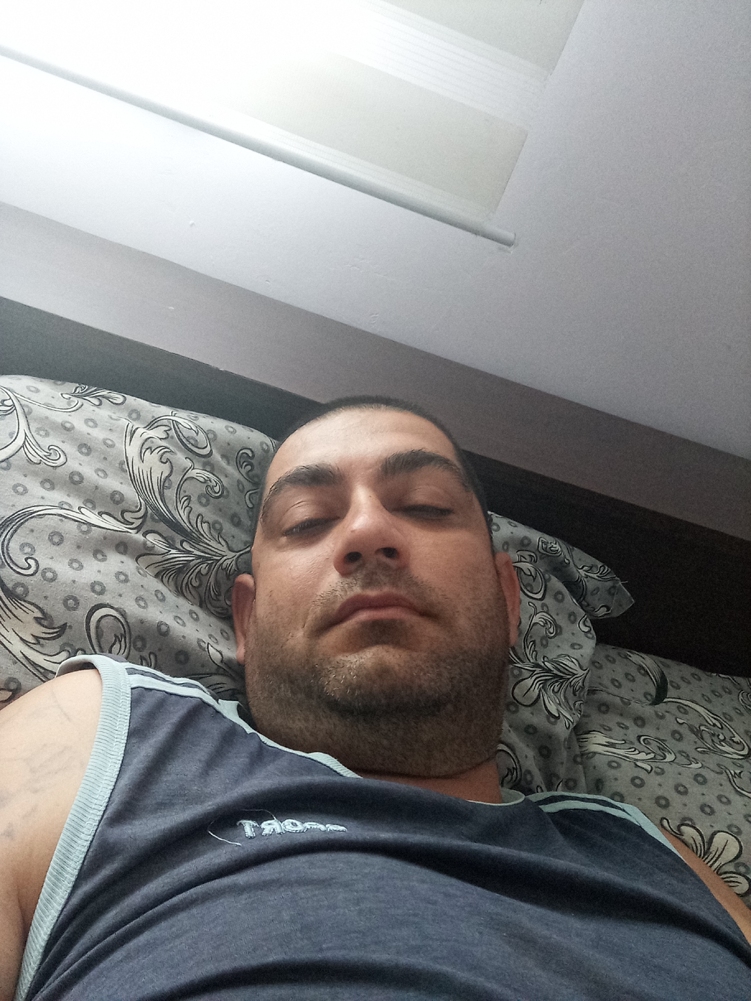 A man is lying on a bed, with his head resting on a pillow with a floral pattern. He is wearing a dark blue tank top. The ceiling is visible above the bed, and a light fixture is mounted on it. The image appears to be taken in a bedroom. The location in Ravnets, Bulgaria is not specified in the image.