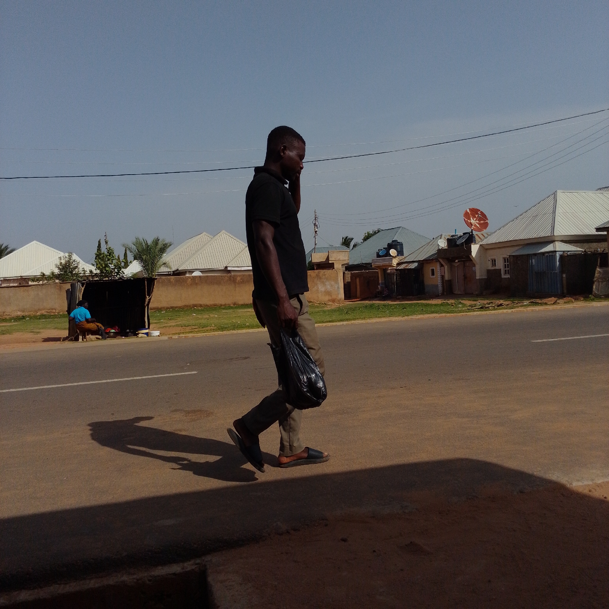In Gombe, Nigeria, a man in a black shirt and tan pants walks along a paved road, holding a black plastic bag in his right hand and talking on his phone. He wears dark sandals. In the background, several houses with metal roofs are visible. To the left, a person in a blue shirt and brown pants sits on a stool in front of a makeshift stall. The sky is clear and blue.