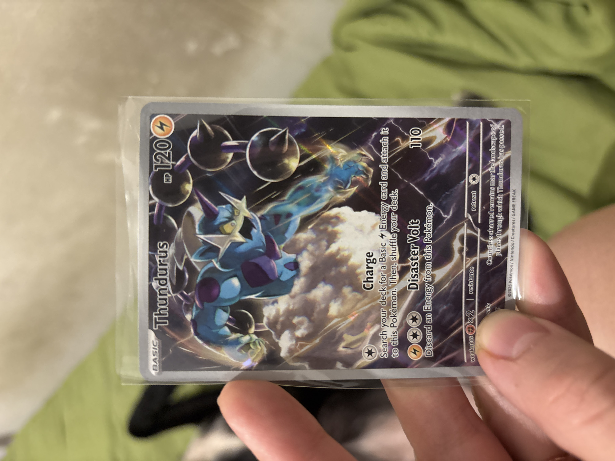 A person is holding a Thundurus Pokémon card in a protective plastic sleeve. The card is predominantly silver and black, featuring an image of the Thundurus Pokémon. The card's text includes the Pokémon's name, "Thundurus", its type, "Basic", its HP "120", and attack descriptions "Charge" and "Disaster Volt". The attack "Disaster Volt" does 110 damage. The bottom of the card has a copyright date of "©2025 Pokémon/Nintendo/Creatures/GAME FREAK". The background of the scene shows a green and crumpled fabric. The location of Buenos Aires, Argentina is not relevant to the image, as the image is a close-up of a Pokémon card.