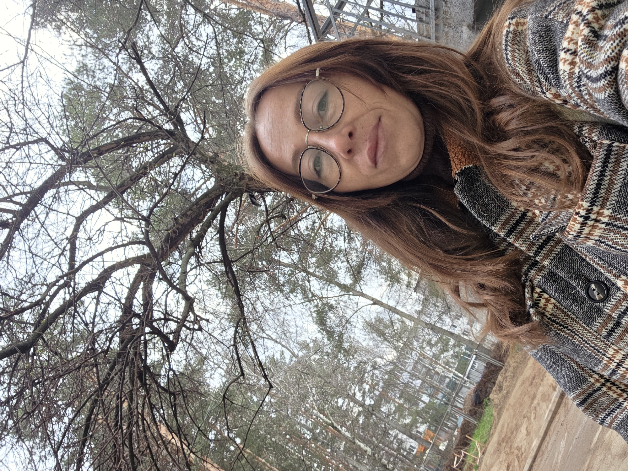 The image is a tilted, low-angle selfie of a woman outdoors in Chaikovsky, Russia. She has long, wavy reddish-brown hair and wears large, round glasses with a tortoise-shell pattern frame. Her expression is neutral as she looks directly at the camera. She is dressed in a dark brown turtleneck top and a patterned jacket featuring a grey, brown, and black plaid design.
The background consists primarily of a dense network of bare tree branches against an overcast, light sky, indicative of late autumn or winter. Some faint green foliage from evergreen trees is also visible. Through the trees in the distance, parts of industrial or residential buildings can be discerned. The lower right section of the image reveals an area of disturbed earth, possibly a construction site or a cleared path, with some faint green vegetation. No text is visible in the frame.