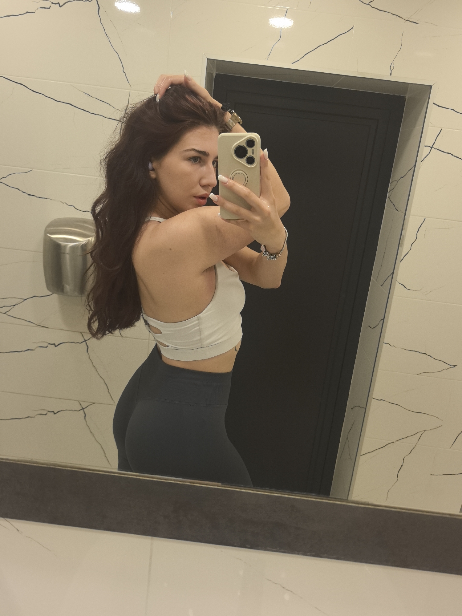 The media file shows a young woman taking a mirror selfie in a modern bathroom. She has long, wavy auburn hair and is wearing a white sports bra and dark gray leggings. Her left hand is touching her hair, and her right hand is holding a smartphone, angled to capture the reflection. She is wearing a watch on her left wrist and a bracelet on her right. The bathroom features white marble-like tiles with black veining on the walls, and a dark gray door is visible in the background. A silver hand dryer is mounted on the wall to the left. The lighting suggests it is daytime. The woman appears confident and posed. There is no visible text in the image. The location context is Stara Zagora, Bulgaria.