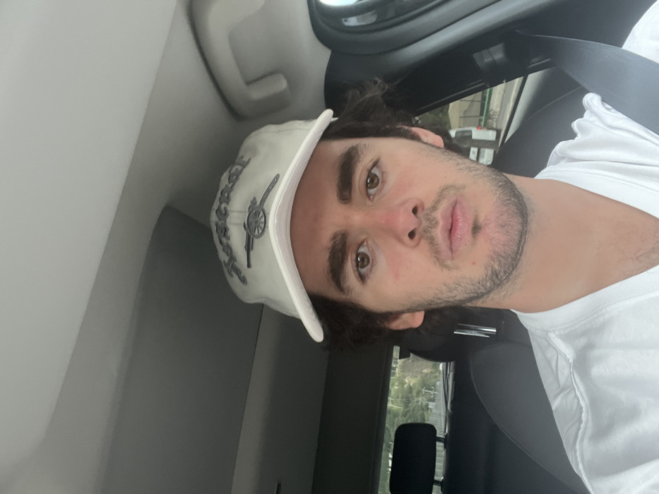 A young adult male is depicted seated in the driver's seat of a vehicle, viewed from the chest up. He has dark, wavy hair, brown eyes, pronounced eyebrows, and light facial stubble, with a neutral expression directed towards the camera. He wears a white short-sleeved t-shirt and a light-colored baseball cap with "Arsenal" in stylized lettering above a cannon emblem. A dark seatbelt diagonally crosses his right shoulder and chest. The vehicle's interior shows a light-colored headliner and a dark-colored seat headrest in the background. Through the rear left window, green foliage and a light-colored pole are visible. Through the front right window, part of a white van or bus is discernible, along with additional green foliage. The scene is located in the city of Unknown, Unknown.